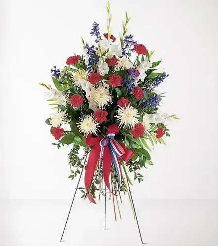  Patriotic Tribute Standing Spray - Honor a life of service and devotion with our Patriotic Tribute Standing Spray. This magnificent arrangement proudly displays the colors of the American flag, blending vibrant red carnations, pure white spider mums, and striking blue delphiniums. Accented with lush greenery and adorned with a classic red, white, and blue ribbon, this standing spray is a heartfelt tribute to those who have served their country or shown unwavering patriotism.  Expertly crafted on a sturdy easel, the Patriotic Tribute Standing Spray stands tall as a symbol of courage, pride, and unwavering dedication. Perfect for military services, veterans, or anyone who lived with a spirit of patriotism, this elegant arrangement brings comfort and honor to any memorial or funeral service.  Let this spray express your deepest respect and gratitude, serving as a beautiful reminder of a life well-lived.