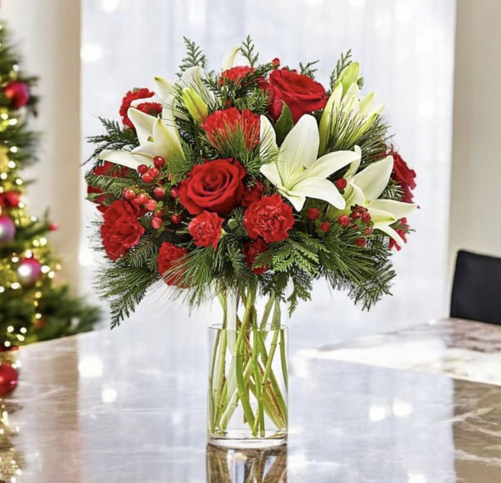 Christmas Snowlight - Bring the magic of the season into any space with the Christmas Snowlight - a festive blend of holiday classics designed to brighten the winter days. Pure white lilies open like soft snowflakes among rich red roses and vibrant red carnations, while clusters of winter berries and fresh evergreen branches add traditional Christmas warmth and texture. Arranged in a glass vase for a clean, timeless look, this bouquet makes the perfect holiday gift or centerpiece. Its fresh seasonal fragrance and striking red-and-white palette create an instant atmosphere of joy, elegance, and celebration.