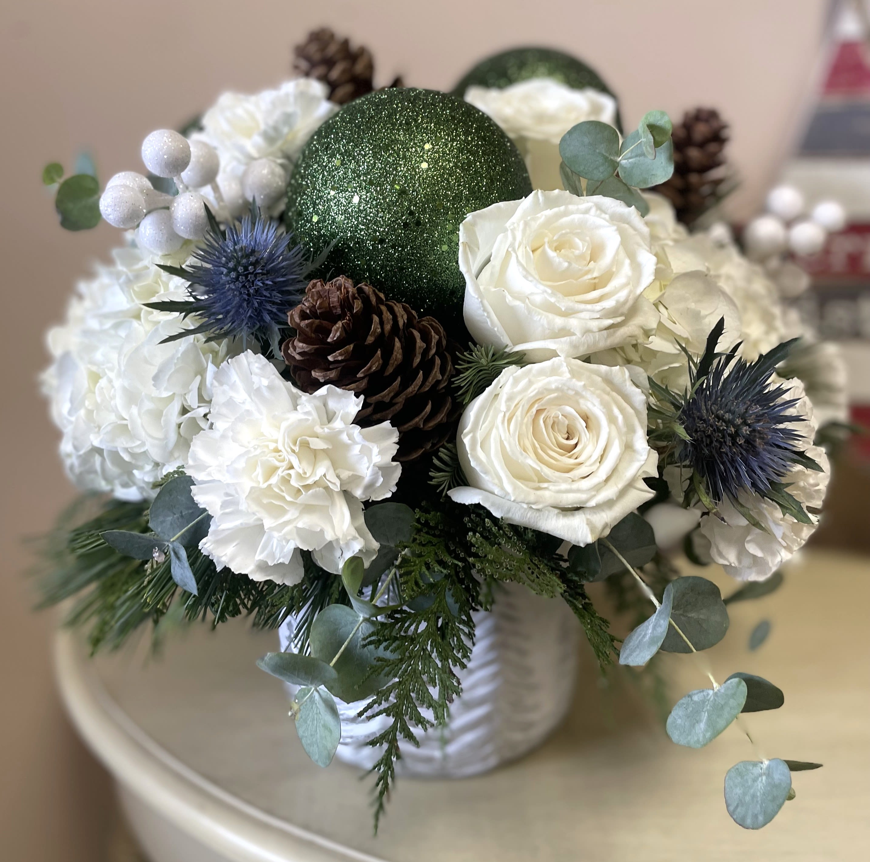 Winter Whites - This elegant arrangement captures the serene beauty of winter in a ceramic pot featuring silver pine trees. Lush wintergreens provide a rich backdrop, accented by soft white hydrangeas, roses, and carnations. Striking blue thistles add a cool contrast, while pinecones bring an earthy, woodland touch. Green ornament balls and silvery eucalyptus add festive sparkle and freshness, creating a sophisticated, seasonal centerpiece that evokes the peaceful charm of a winter landscape.