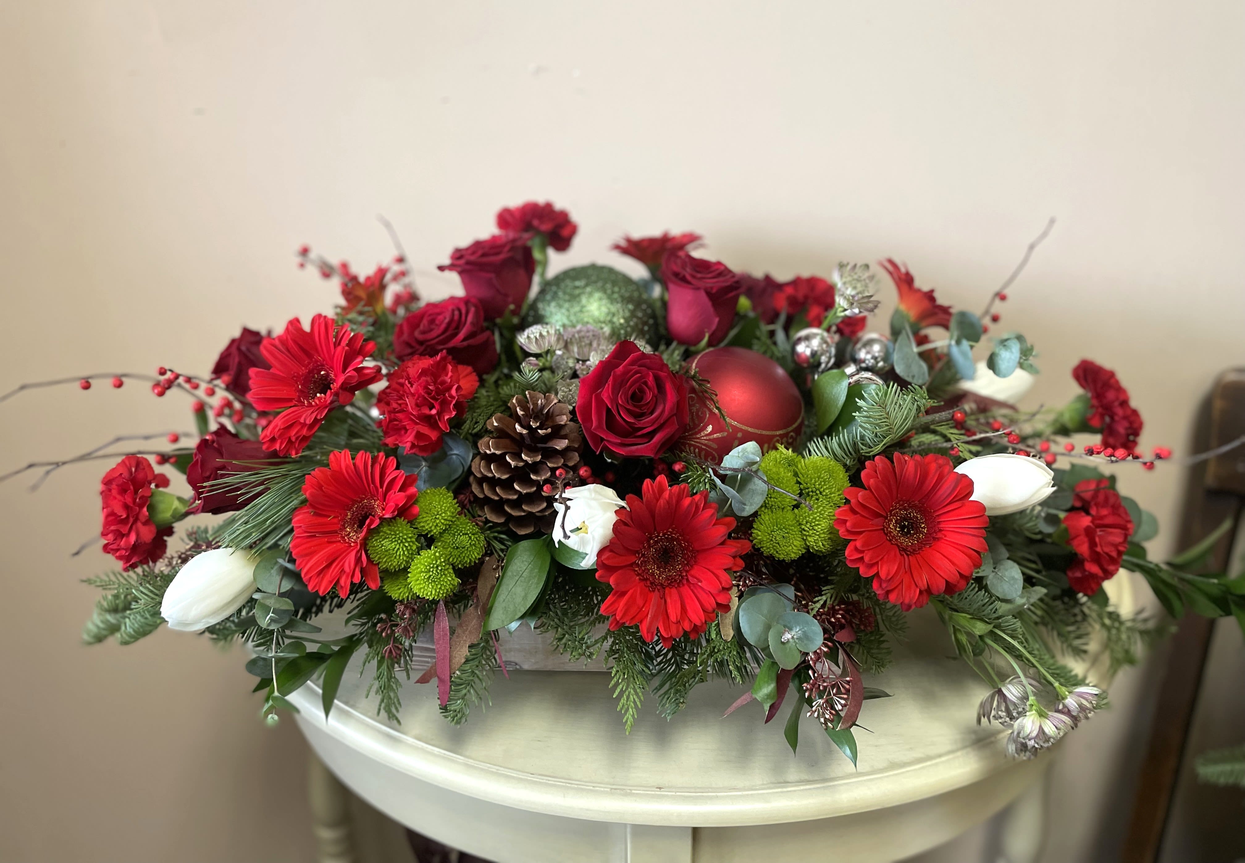 Merry &amp; Bright - Long and low arrangement set in a charming vintage wooden box, evoking a rustic yet festive feel. It features an assortment of winter greens, including sprigs of pine and cedar, creating a lush, fresh foundation. Red roses, with their velvety petals, are nestled alongside rich red carnations, both adding a deep, romantic touch. Bright lime green mums provide a vibrant contrast, their spiky petals bringing a playful pop of color to the arrangement.  Scattered throughout are festive ornaments, gleaming in metallic red, green and silver catching the light and enhancing the holiday spirit. Sprigs of winterberry with their bright red berries weave through the greens, adding a touch of nature’s winter charm. Red gerbera daisies, with their striking, large petals, bring a cheerful brightness to the composition. Pine cones, dusted with a light touch of faux snow, add texture and an earthy feel, completing the look with a cozy, seasonal touch. The overall design is rich, textured, and joyful—perfect for a holiday table or mantel.