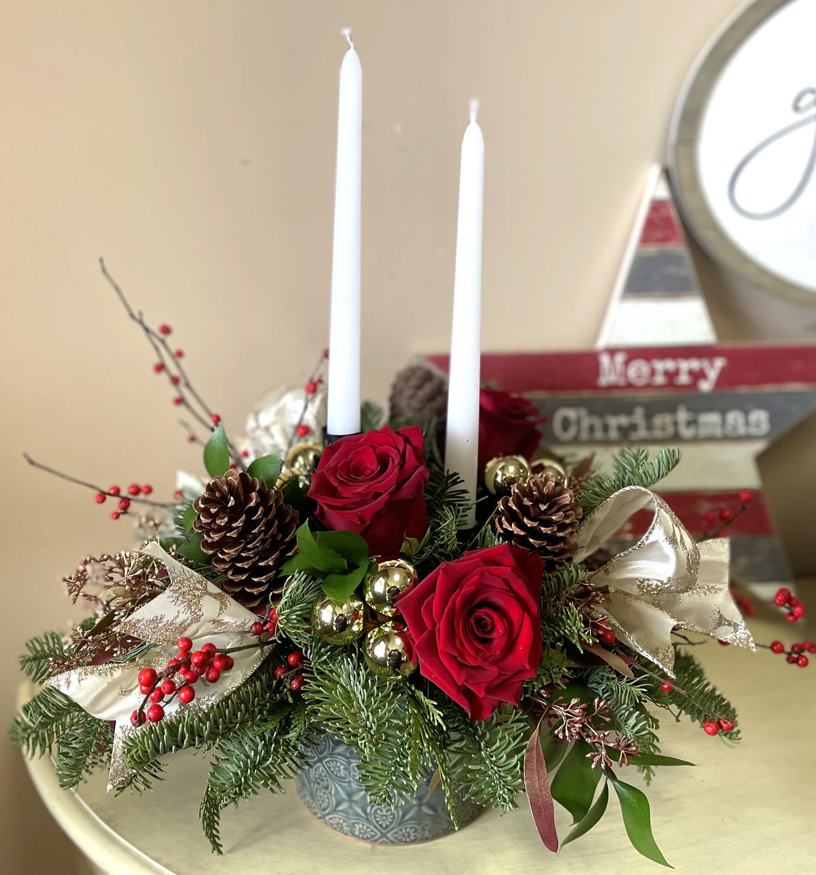 Willow Winter - This winter flower arrangement combines vibrant red roses, bright red winter berries, and lush green pine and cedar sprigs, evoking the beauty of the season. Pine cones add a rustic touch, while two white candles provide a soft, glowing warmth. All of this is elegantly arranged in a deep blue ceramic container, creating a festive and serene centerpiece perfect for the holidays.