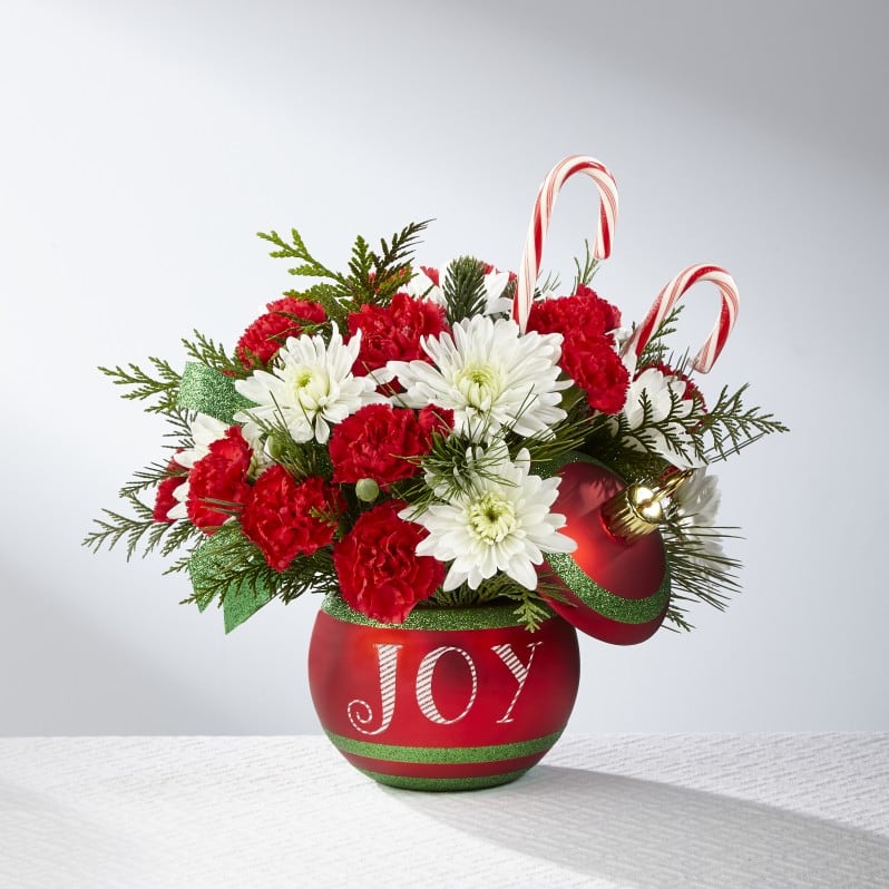 The FTD® Season's Greetings™ Bouquet: 17-C5s - The FTD® Season's Greetings™ Bouquet is ready to dazzle your special recipient this holiday season! Rich red roses, red mini carnations, white carnations, and white chrysanthemums, accented with fragrant holiday greens are arranged by our floral artisans to create a very merry Christmas bouquet. Presented in a keepsake sparkling red ceramic ornament inspired vase, accented with green stripes and the word, "Joy," on the front in white, with a matching lid to create a treasured Christmas accent piece, this holiday bouquet is also embellished with two peppermint candy canes to create a truly remarkable Christmas gift.