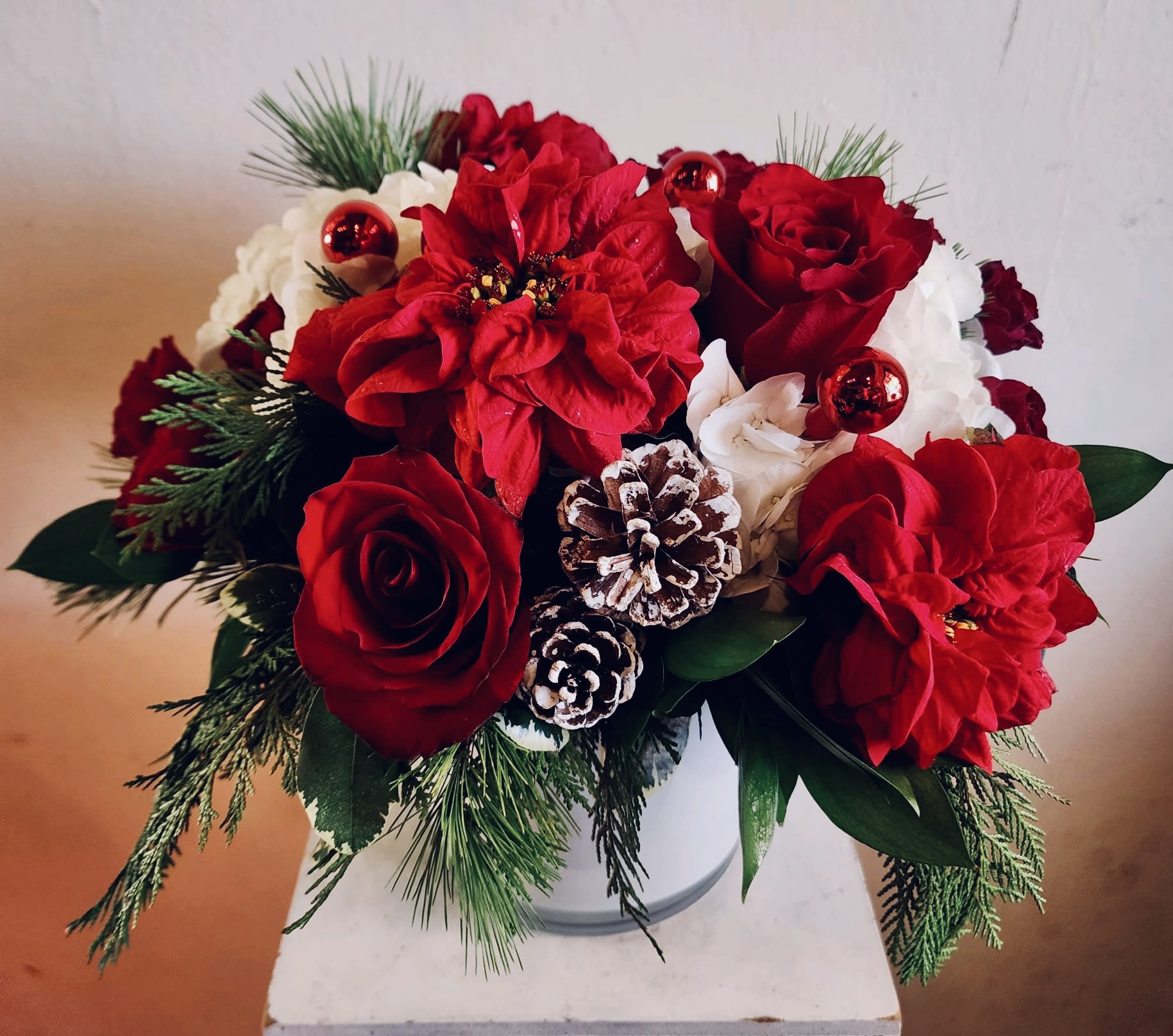 Deck the Hall - White hydrangea, red roses, winter rose poinsettias, accents of ornaments and pine cones with mixed winter greenery. White or leaf ribbon-wrapped clear vase upon availability 