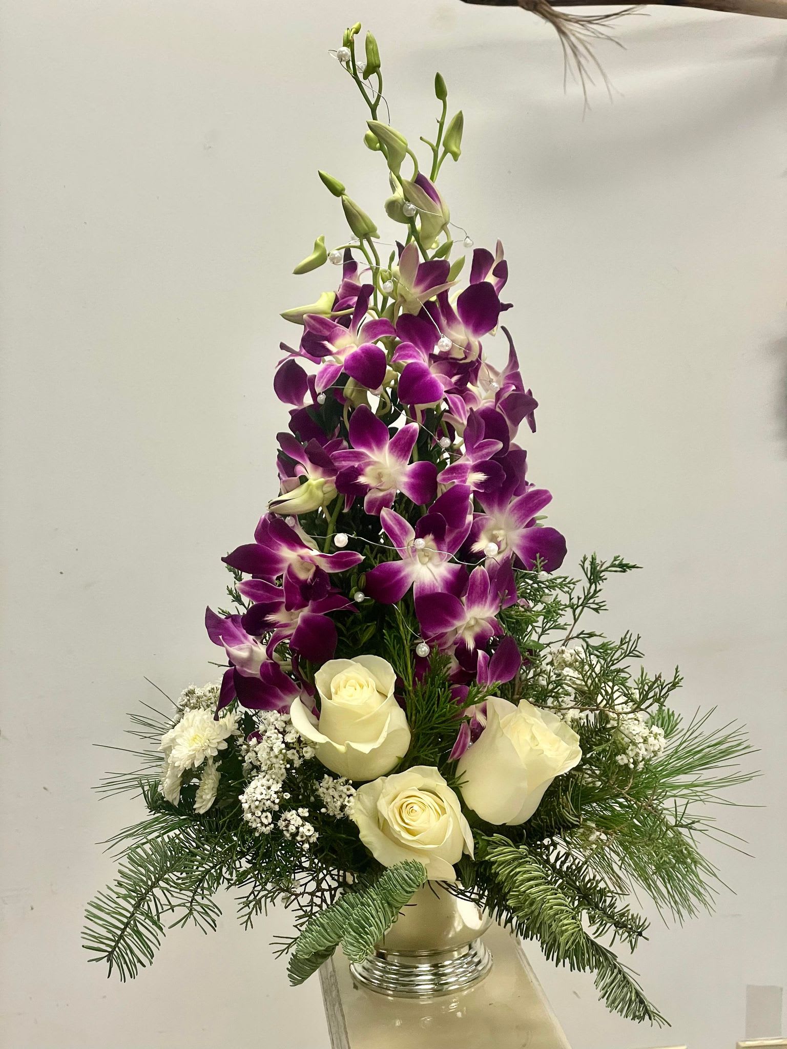 Festive Orchid Elegance - Celebrate the holiday season in exquisite style with our enchanting Festive Orchid Elegance arrangement. This stunning floral masterpiece captures the essence of Christmas in a unique and sophisticated display. Nestled in a short pedestal silver container, the arrangement takes on the form of a graceful Christmas tree, instantly infusing any space with seasonal joy.