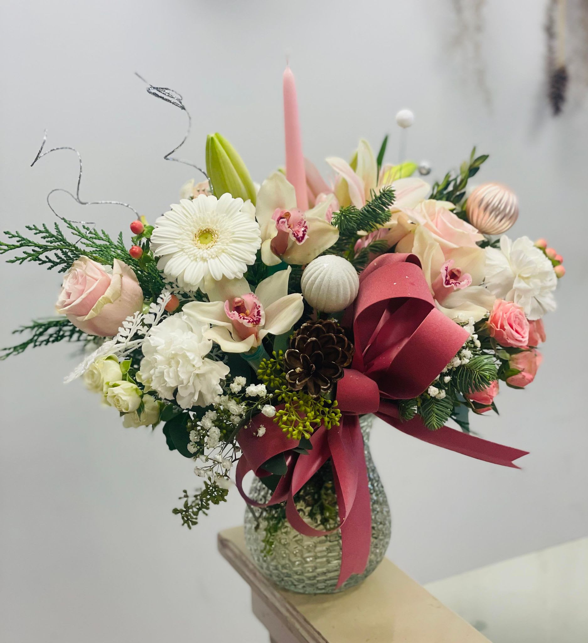 WARM GLOW - This winter holiday flower arrangement is a celebration of contrasts, blending the purity of white and pink with the opulence of silver, the rustic charm of pinecones, and the timeless allure of Christmas ornaments. It is a masterpiece that captures the magic and joy of the season, a perfect centerpiece to grace your festive gatherings and infuse your home with the spirit of winter enchantment.      