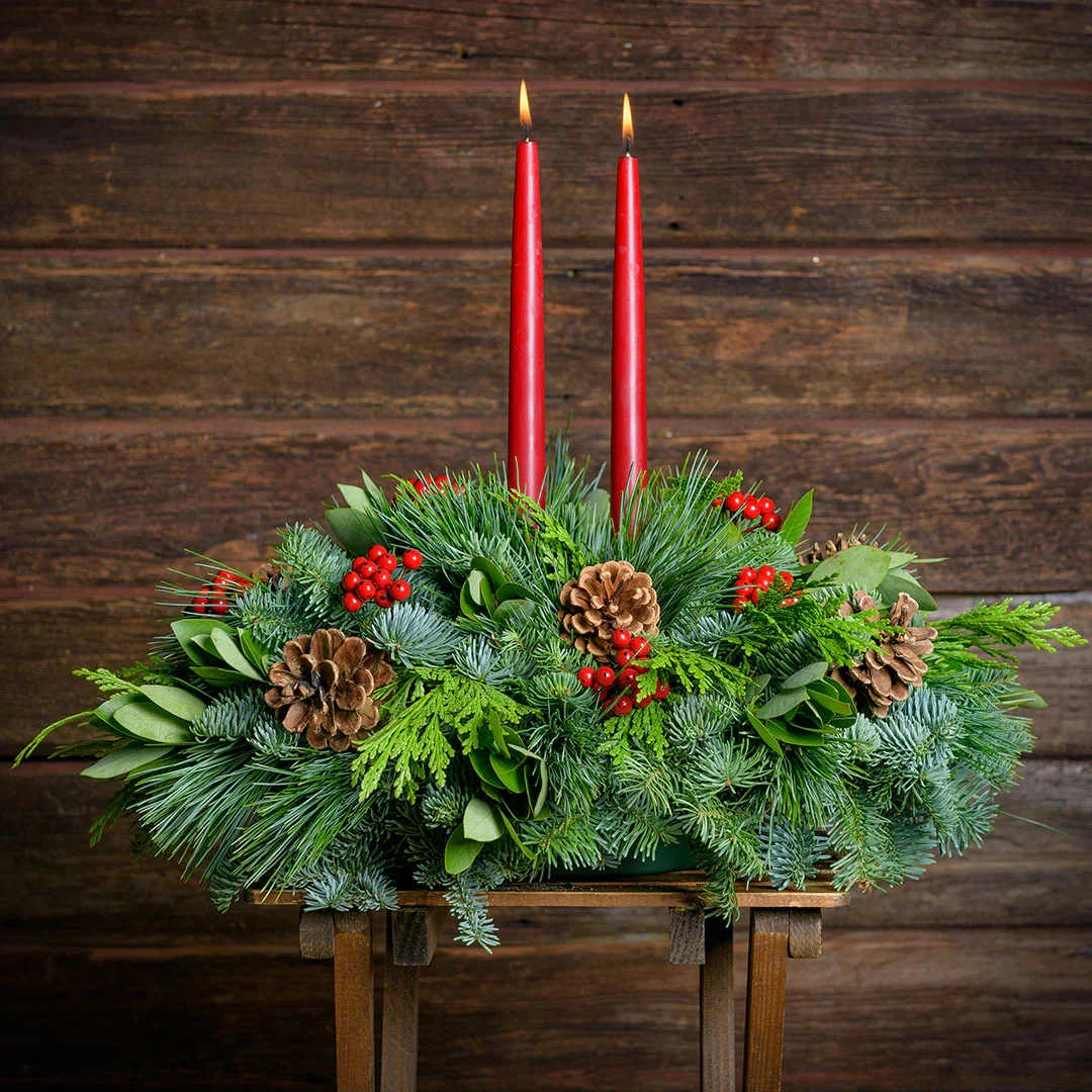 Red Pacific Bay Centerpiece - Over the years, Centerpiece has become a Christmas favorite and one of our best-selling holiday centerpieces. This traditional centerpiece arrangement combines a delightful blend of fresh noble fir, cedar, and pine with beautiful green bay leaves. Two taper candles light up the centerpiece while ponderosa pinecones and faux berries add the perfect touches. This lovely classic is sized at approximately 14&quot; x 18&quot;. Available in red.