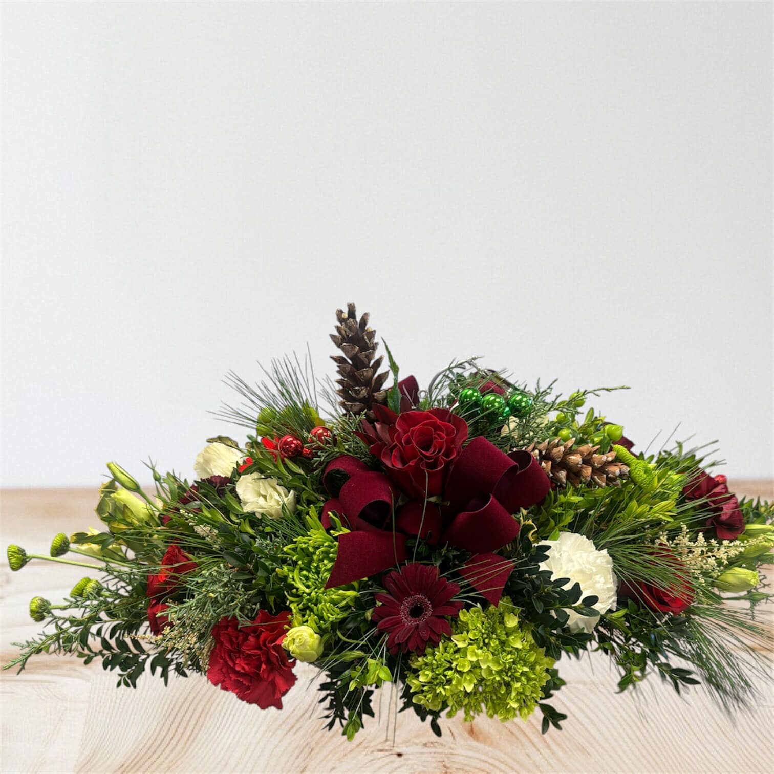 Holiday Cheer Centerpiece - Add a touch of festive elegance to your table this season! This stunning arrangement features rich red roses, carnations, and gerbera daisies complemented by fresh greenery, pinecones, and delicate white blooms. Finished with holiday accents and a deep red bow, it’s perfect for Christmas dinners, holiday parties, or as a thoughtful gift.