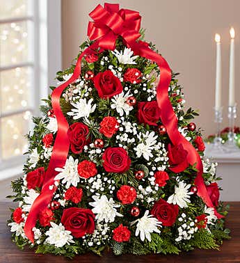 Holiday Flower Tree® With Ribbon - The holiday favorite that started it all! Our flower tree arrangement is popular for its festive style and compact size, which fits perfectly into any space. Gathered with fragrant evergreens, then trimmed with decorative ornaments and a merry red bow on top, this floral twist on a traditional tree makes a truly original gift for family, friends, or your own home.  Tree-shaped arrangement with red roses and mini carnations; white cushion poms; accented with baby’s breath, assorted Christmas greenery, red ball ornaments and a red bow topper
