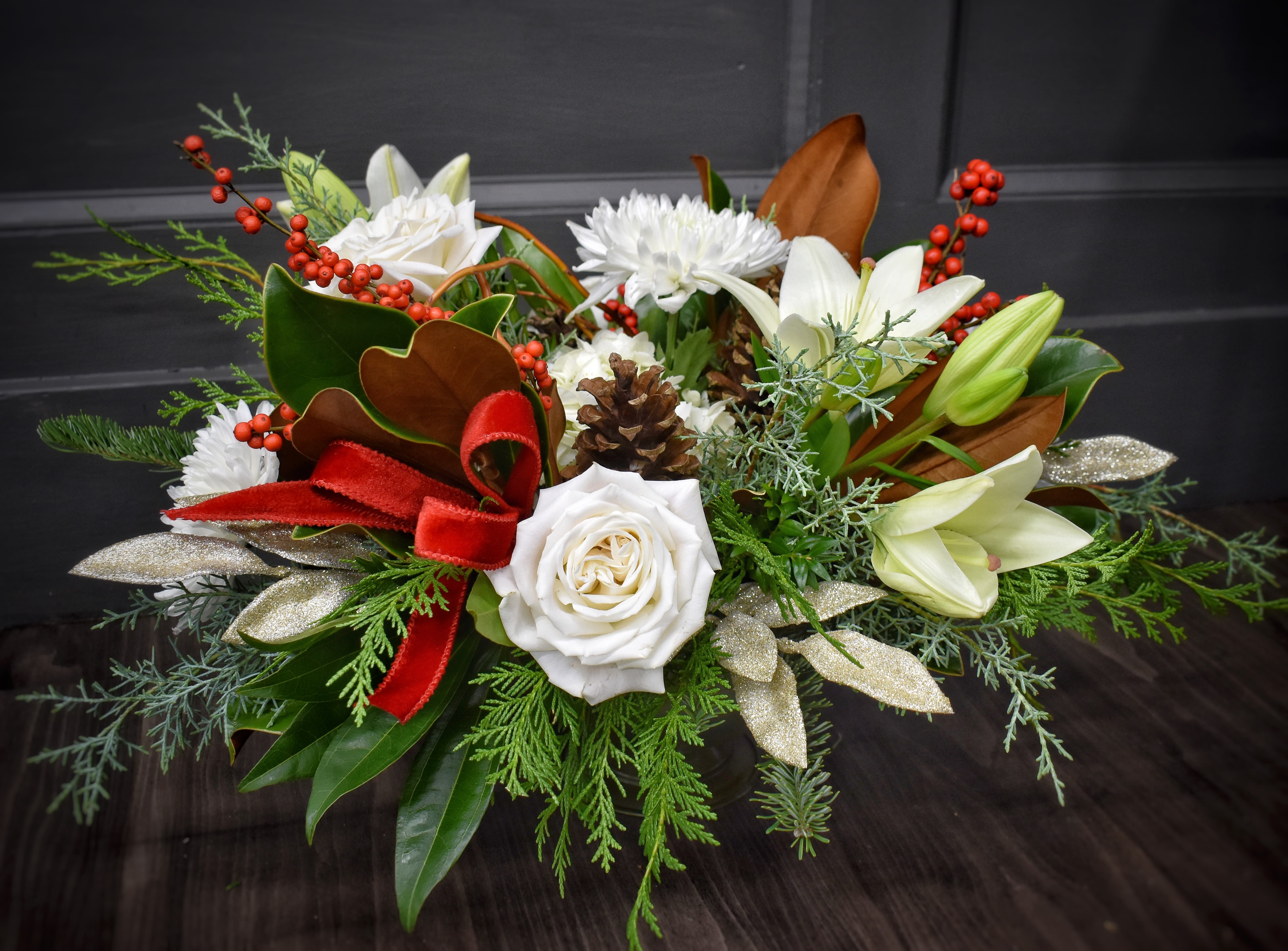 Evergreen Elegance - Our biggest and classiest winter centerpiece of the year! This luxurious design is arranged in a large footed container and features magnolia and evergreen foliage, pine cones, large focal flowers including hydrangea, lilies, roses, and cremon mums accented with gold silk leaves and red velvet ribbon. 