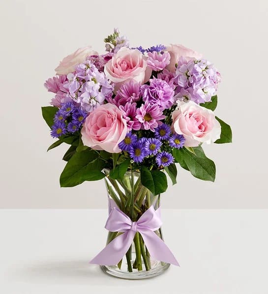 Dreamy Day Bouquet - Like something out of a dream, this enchanting lavender bouquet is artfully arranged with soft pink accents and lush greenery. Presented in our elegant fluted violet vase—crafted from vintage-inspired pressed glass with delicate ribbed detailing—it beautifully showcases the rich hues of the flowers and the heartfelt sentiment behind your gift.  All-around arrangement with pink roses; lavender carnations, daisy poms, button poms and stock; purple monte casino; accented with assorted greenery Artistically designed in our exclusive fluted gathering vase in a lovely shade of violet; vintage pressed glass with ribbed detail creates a charming enhancement for the floral arrangement; measures 7.9"H x 5"D Extra large arrangement measures approximately 17"H x 14"W Large arrangement measures approximately 15"H x 13"W Medium arrangement measures approximately 14"H x 12"W Small arrangement does not include monte casino; measures approximately 13"H x 10"W Our florists hand-design each arrangement, so colors and varieties may vary due to local availability