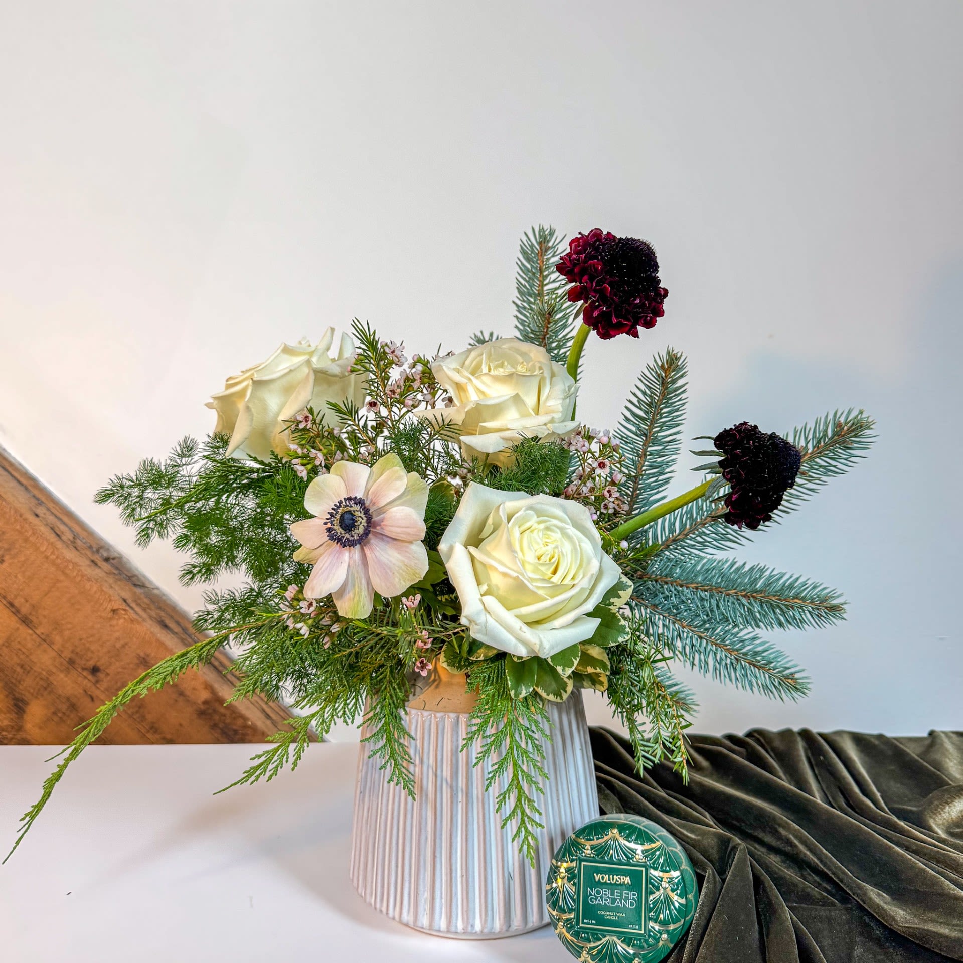 Noble Fir Gift Set - Elegant and fragrant, this set brings the fresh serenity of a winter forest indoors. Includes a fresh neutral arrangement in a ceramic vessel, and a 4 oz voluspa tin candle in the scent "Noble Fir Garland". Arrangement measures approx 9" x 12". Upgrade to deluxe or premium for additional luxe blooms. 