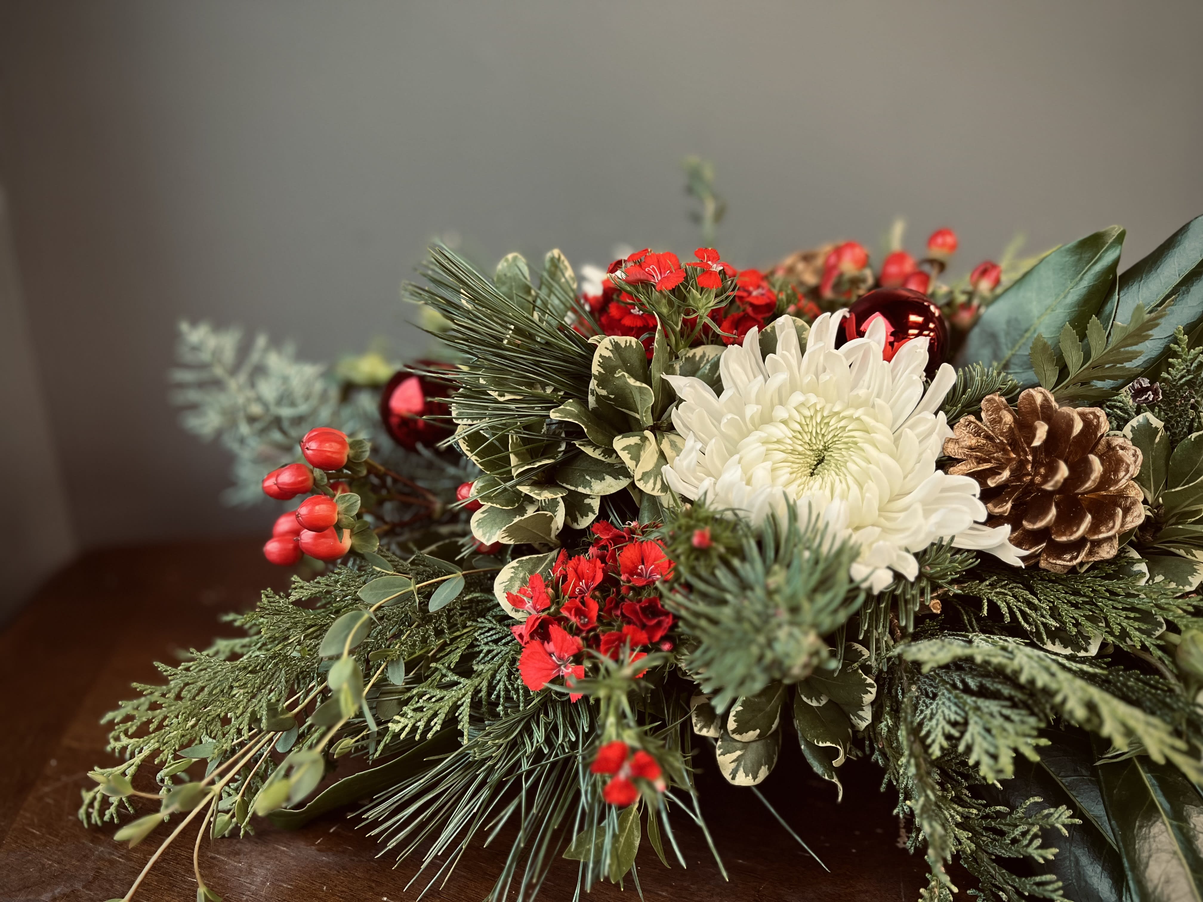 Holiday Greetings Centerpiece  - Spread joy at your holiday gathering this year with this traditional array of wintergreens with pops of white and red florals. Designed in a no-show container, this arrangement is the perfect accent for the dinner table. Upgrading to to deluxe or premium will allow us more budget towards florals.  