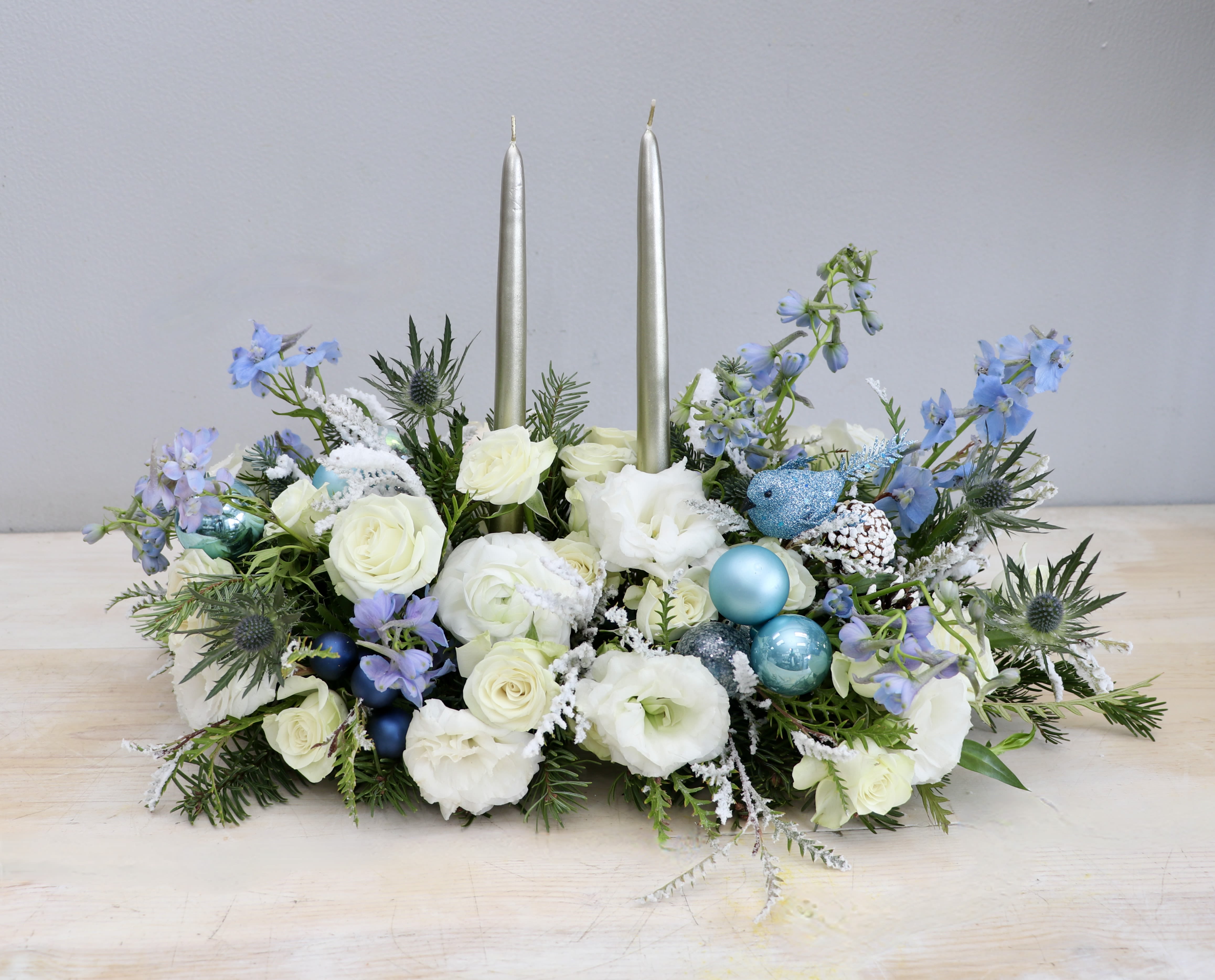 Celebrate in Blue - WeHo Florist - This holiday centerpiece is made with white florals with touches of blue delphinium and thistle. Accented with fragrant greens and blue ornaments, this centerpiece is perfect for any holiday celebration! Standard is about 12"