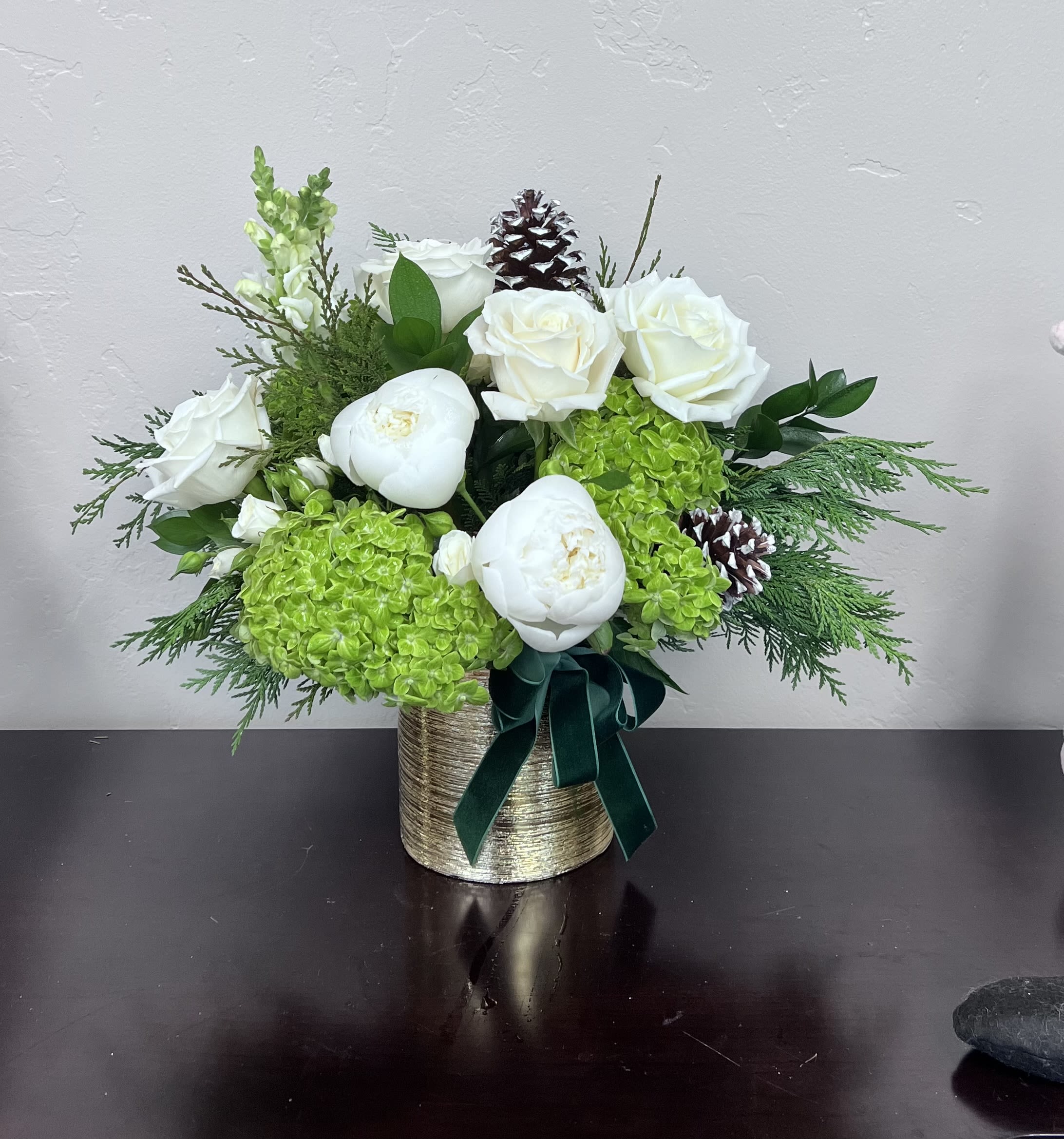 Snowfall In The Pines Christmas Floral - A breathtaking green-and-white holiday arrangement featuring lush green hydrangea, pristine white roses and peonies, delicate white snapdragons, princess roses, and beautifully scented evergreens. Rich magnolia foliage evokes cherished winter memories, while natural pine cones and a gold vessel add a touch of festive luxury. An elegant, timeless floral piece perfect for any space.