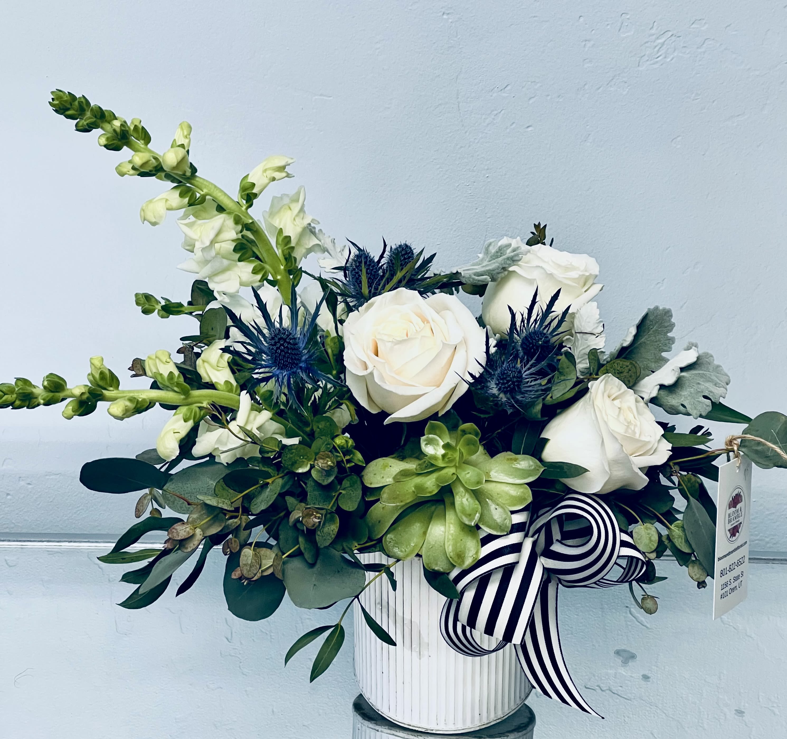 Mountain Blooms -  The Mountain Blossom Arrangement  Soft, romantic, and effortlessly elegant—this design overflows with creamy whites, silvery textures, and lush greenery. Nestled within is a succulent you can keep and grow forever, making this piece as lasting as it is beautiful. Arranged in a vintage-inspired vessel and finished with a striped ribbon, the Mountain Blossom Arrangement brings together rustic charm and feminine grace. A lush floral that feels both timeless and unforgettable. 