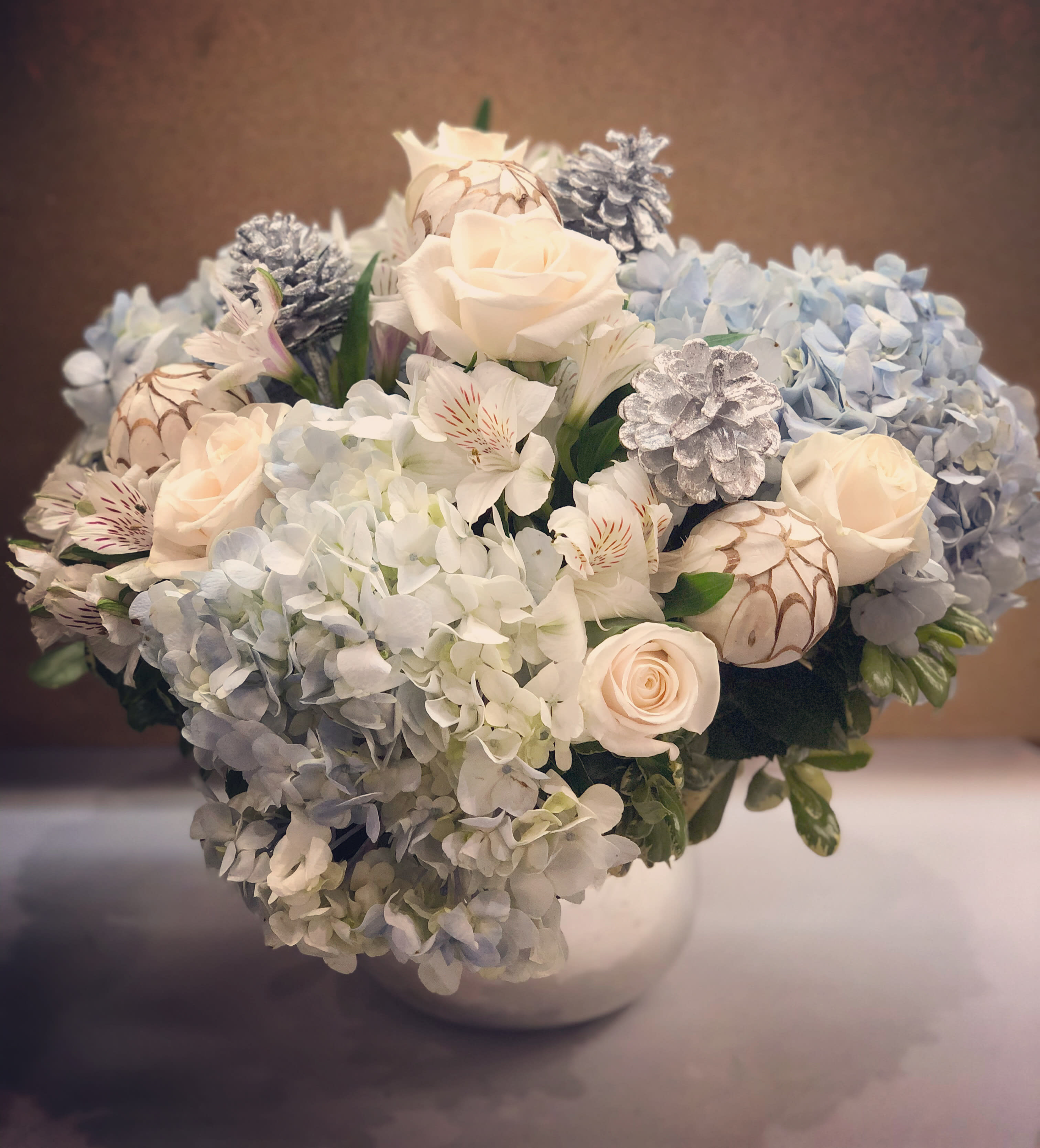 Winter Blues  - A beautiful design of lush seasonal white floral blooms, with light blue accents accompanied by silver and white ornaments and designed in our signature metallic silver vase.                         Orientation: All-Around All prices in USD ($) Standard, Deluxe, Premium (SHOWN)
