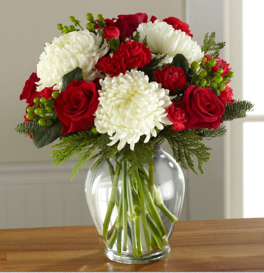 Holiday Enchantment Bouquet - Beautiful, bright, and blooming with a broad holiday appeal, this gorgeous Christmas flower bouquet is simply enchanting at every turn. Gorgeous and full white snowball chrysanthemums are not to be upstaged when arranged amongst rich red roses, red carnations, and red mini carnations, accented with red hypericum berries and assorted Christmas greens to create a standout yuletide display. Presented in an attractive clear glass vase, this gift of flower is ready to create a treasured holiday moment for your recipient.