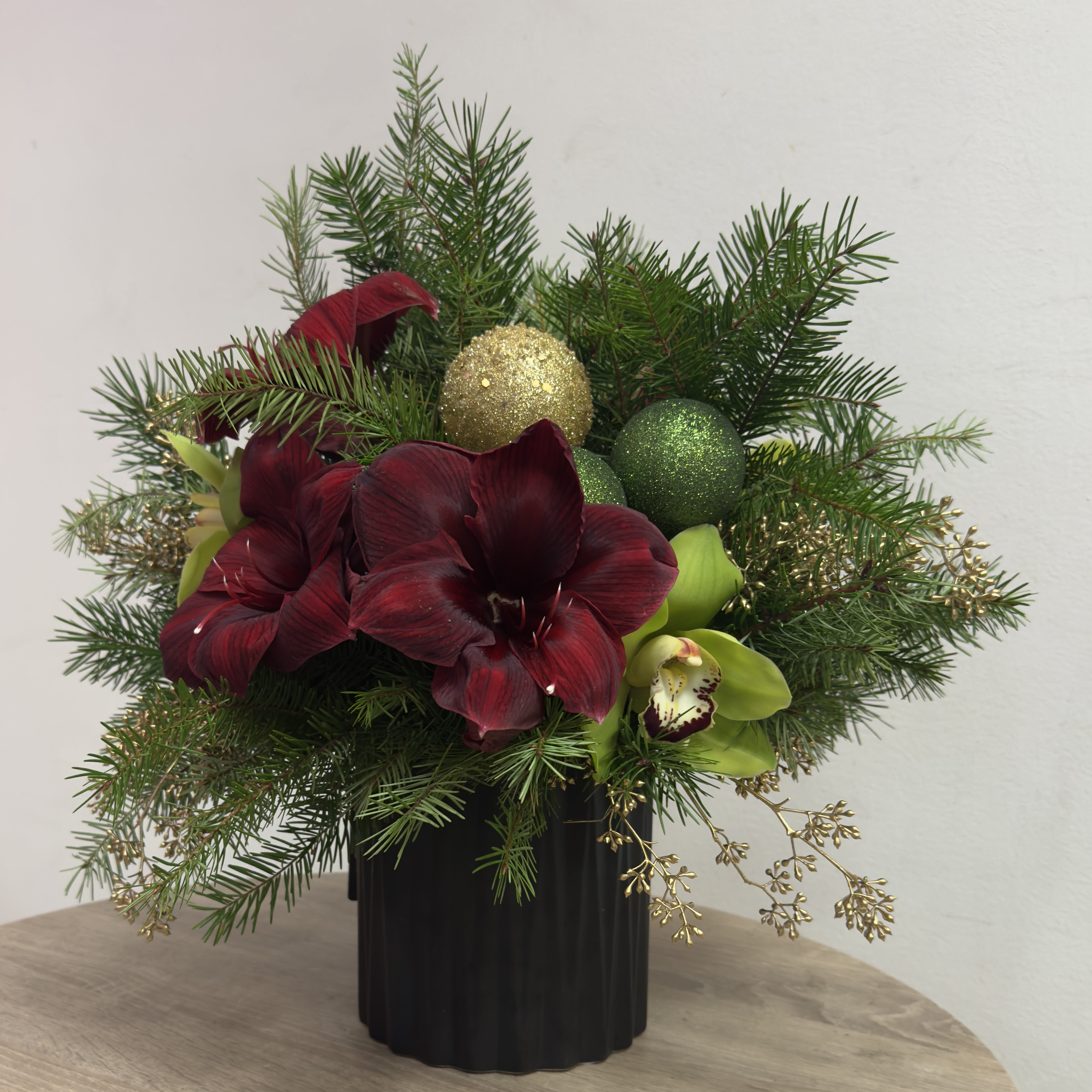 Evergreen Shimmer  - This festive arrangement features deep red amaryllis blooms paired with vibrant green cymbidium orchids, lush evergreen foliage, and sparkling gold and green holiday ornaments. Designed in a sleek black ribbed vase, it’s perfect for adding festive style to any space. Dimensions: Approximately 14" (H) x 12" (W).