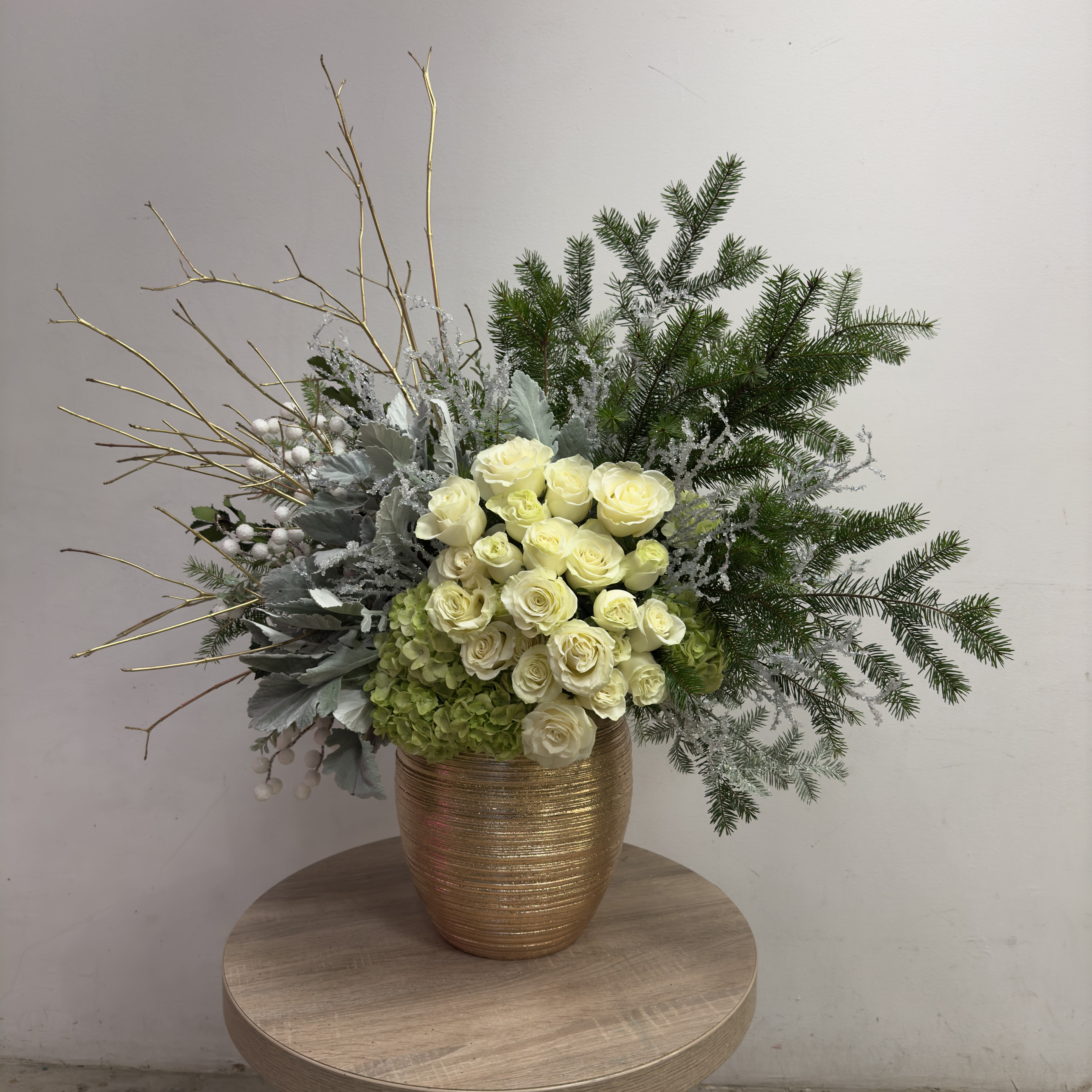 Golden Frost - A luxurious winter-inspired floral arrangement featuring creamy white roses, soft hydrangeas, and lush seasonal greenery, accented with gold branches and frosted elements for a festive touch. Presented in an elegant textured gold vase, this design exudes timeless style.  Dimensions: Approximately 22" (H) x 20" (W) x 12" (D)