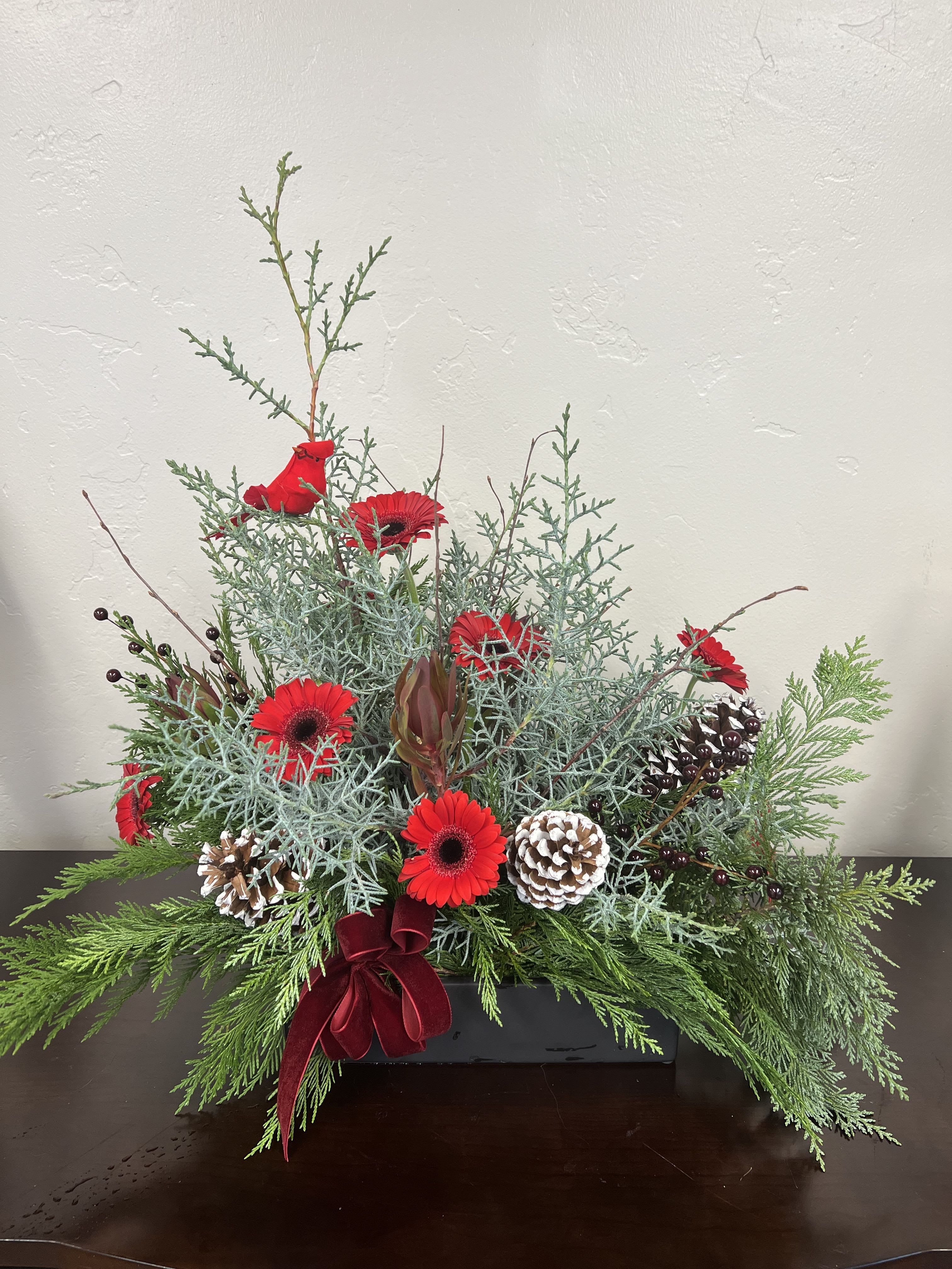 The Woodland Red Robin - The Woodland Red Robin is a festive ode to the magic of the season—rich, fragrant, and full of winter charm. Small Pine Tree, Cedar, and natural Pine Cones bring the unmistakable scents of Christmas indoors, while premium Red Gerberas and Safari Sunset add a warm, luxurious pop of holiday color. Birch twigs, a velvet ribbon, and a charming Red Robin accent complete this artful design. Expertly arranged in a reusable ceramic vessel, this piece is crafted to last. Once the fresh blooms fade, simply remove the flowers and enjoy the evergreens, cones, and textural elements all the way through the holidays with proper watering. A beautiful, long-lasting holiday arrangement—perfect for gifting or elevating your own home décor.