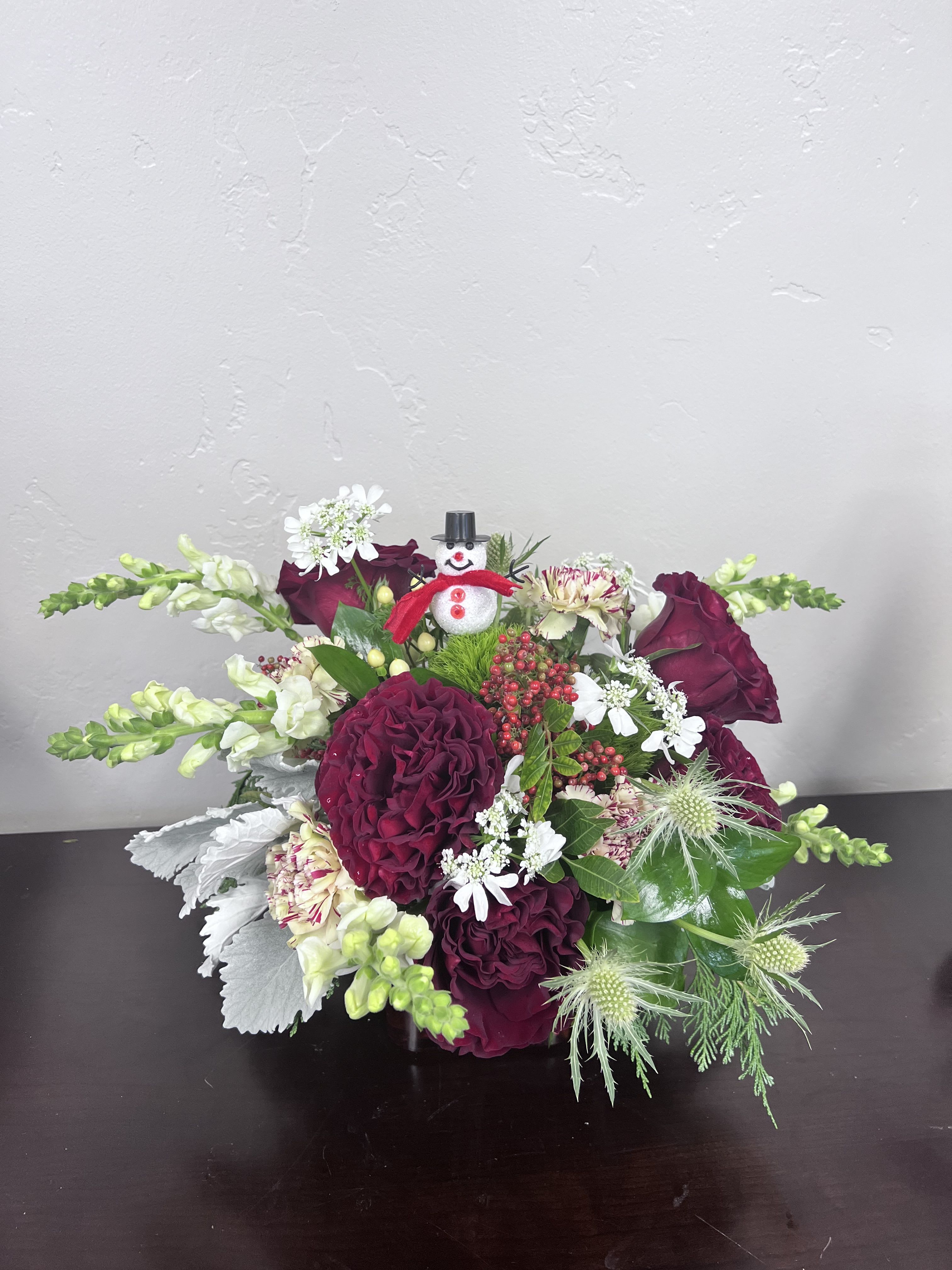 Frosty's Winter Joy - Celebrate the magic of winter with Frosty’s Winter Joy, a cheerful holiday arrangement designed to make every heart—young and old—smile. Lush Red Roses, playful Snapdragons, winter Berries, Carnations, airy Orlaya, and textured Green Thistle come together with aromatic Christmas Greens to create an enchanting ode to everyone’s favorite snowman. Brimming with warmth, color, and holiday charm, this whimsical design brings joy to any space and is sure to delight the kids while adding festive cheer to your home. A perfect seasonal gift that captures the wonder of winter. Comes in a red glass vessel.