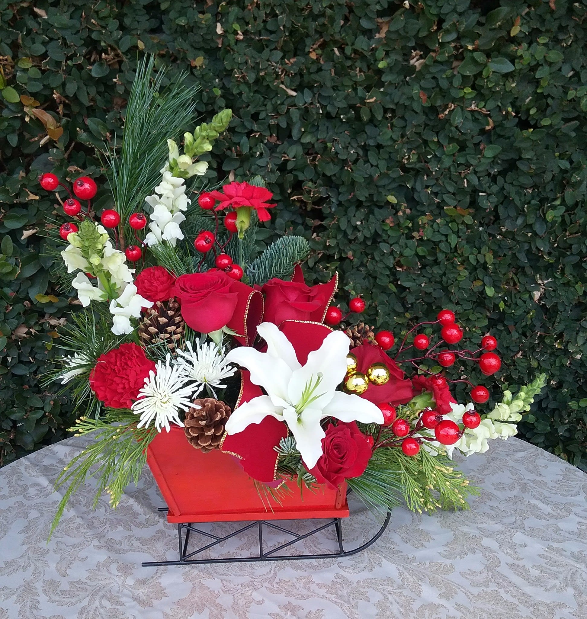 Red Sleigh - White Snapdragons, red roses, red carnations, white lily, white chrysanthemums, pine cones, red glass balls and berries designed in a keepsake wooden Red Sleigh