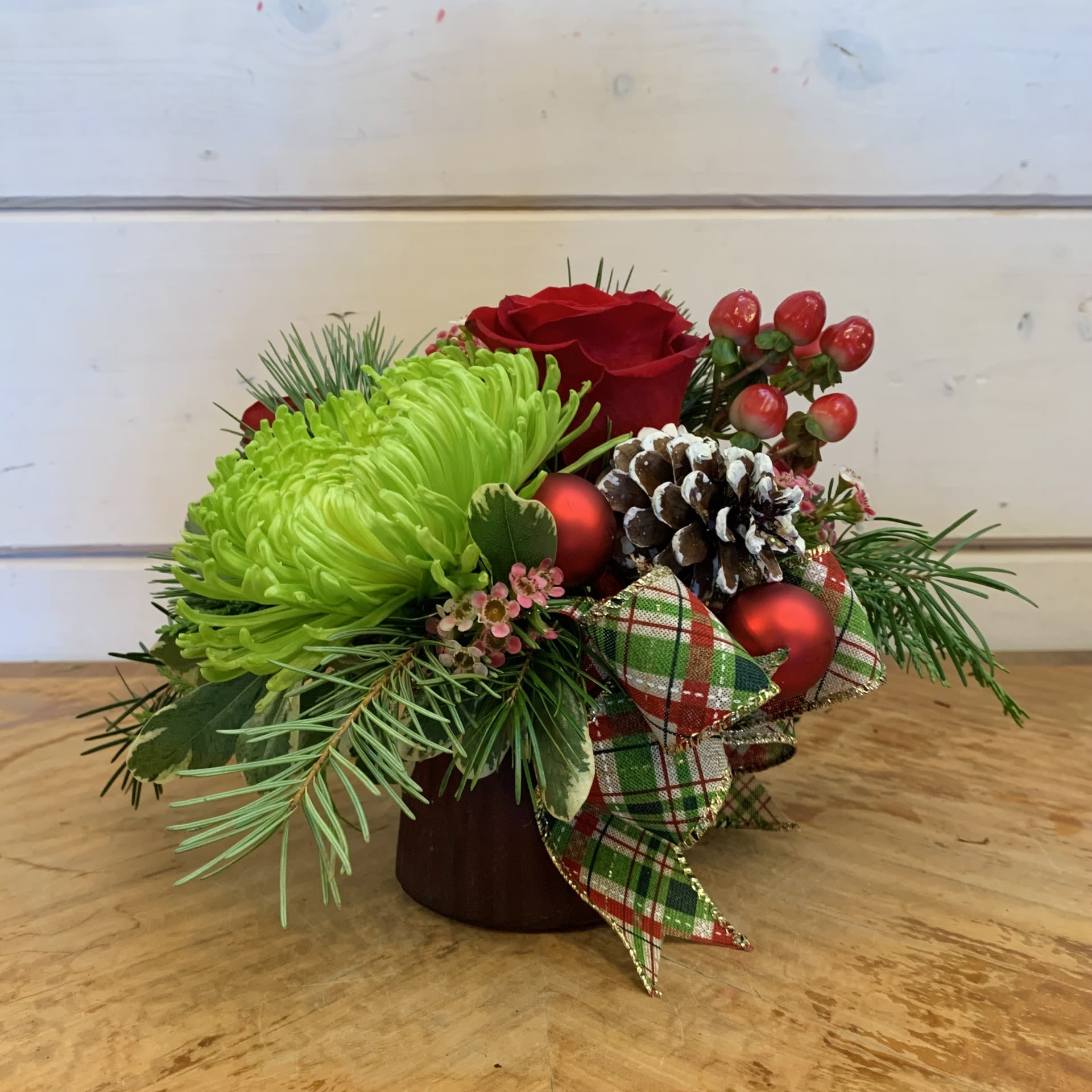 A Little Christmas - A petite classic Christmas arrangement with red roses, green cremon chrysanthemum, red hypericum berries and seasonal wax. Arranged in a petite burgundy glass vase with a plaid bow, red matte ornament balls, and a pinecone. Approximately 7" tall by 7"across.