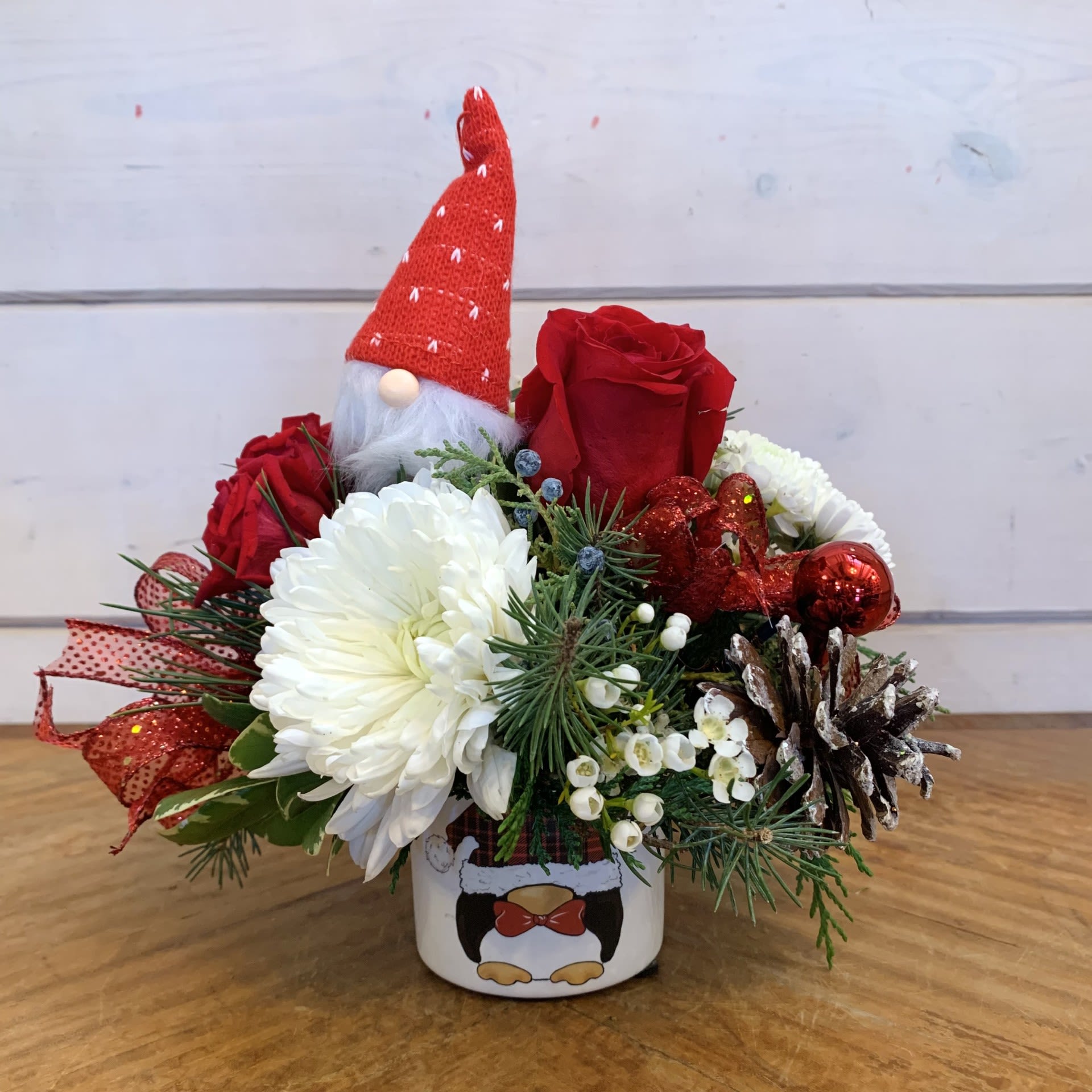 A Little Gnome - This petite arrangement features a petite gnome friend sitting with red roses, red spray roses, and white chrysanthemums in a penguin gnome printed pot with seasonal filler flower, winter foliage, red sparkle bows, shiny red ornament balls and iridescent snowy pinecones. Approximately 7&quot; by 7&quot; for the standard.