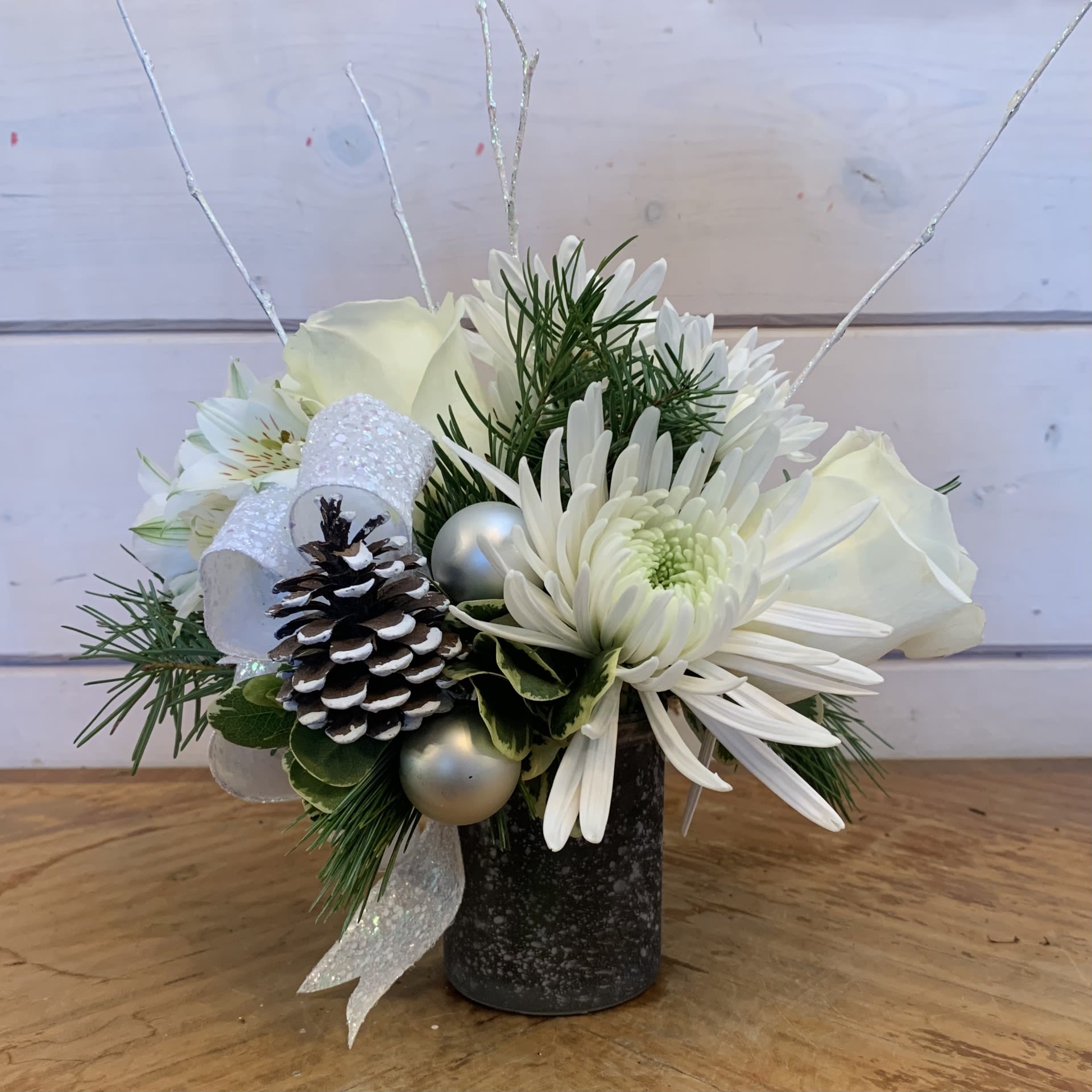 A Little Sparkle - A petite all white winter arrangement with white roses, white cremon crysanthemum, white cushion crysanthemums and white alstroemeria in a frosted silver petite vase with a iridescent bow, white ornament balls, iridescent glitter branches and a snowy pine cone. Approximately 8" across and 8" high (not including glitter branches).