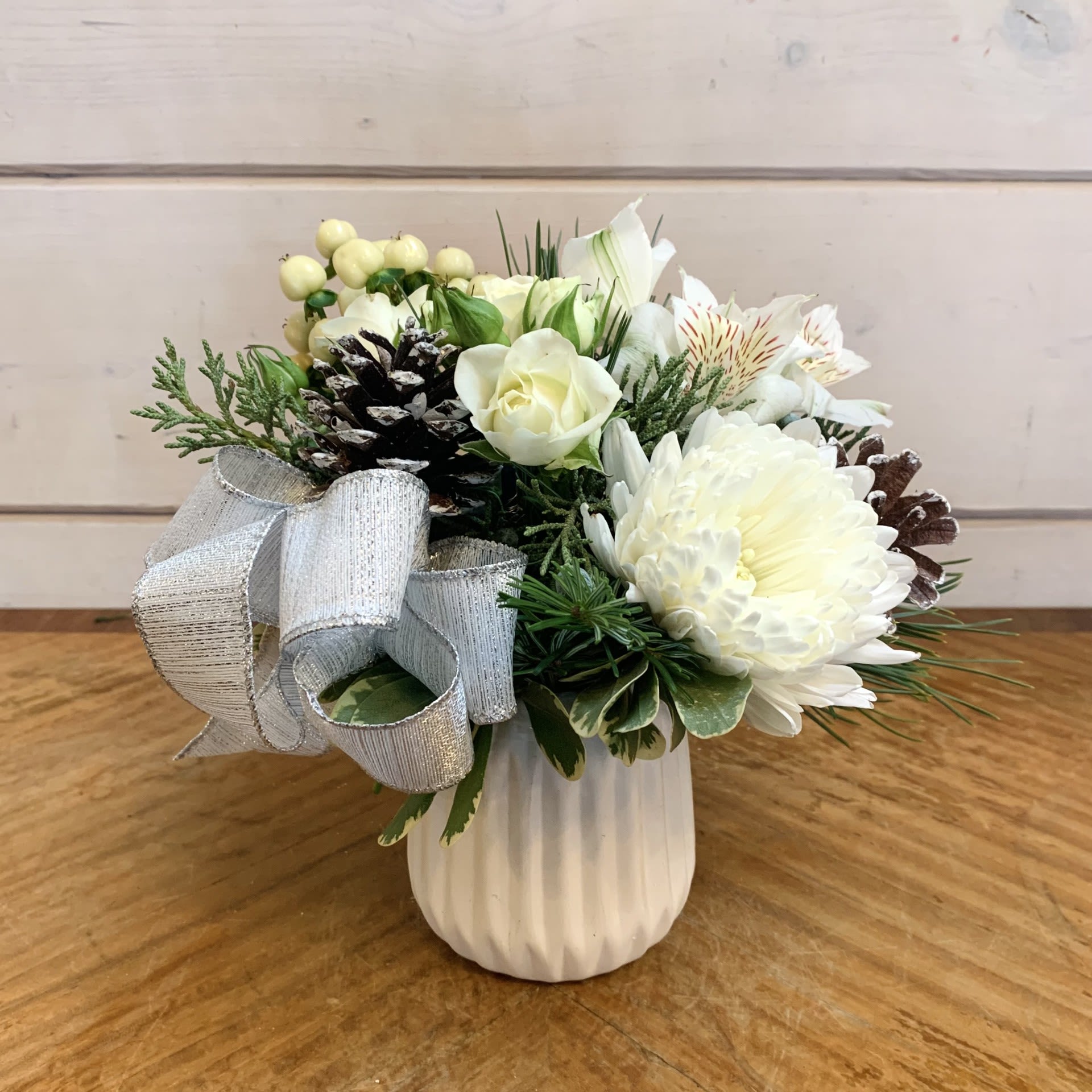 A Little Winter - A petite arrangement of white spray roses, a white cremon crysanthemum, white hypericum and white alstroemeria in a petite patterned white chalk pot with a silver bow and snowy iridescent pinecones. Approximately 6&quot; tall and 6&quot; wide