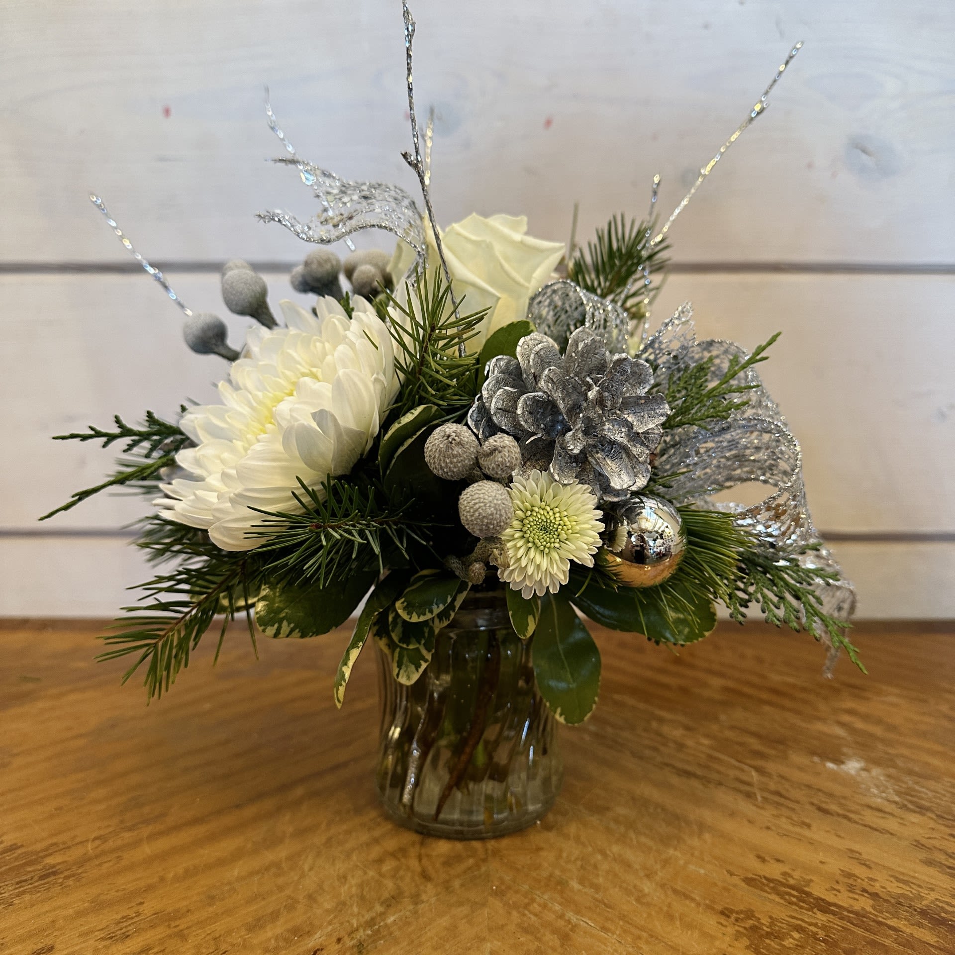 A Little Silver - A petite arrangement featuring white roses, brunia berries, and white chrysanthemums in a petite glass vase with conifer greens, a silver bow, silver pinecones, silver ornament balls, and silver branches. Approximately 6" by 6"