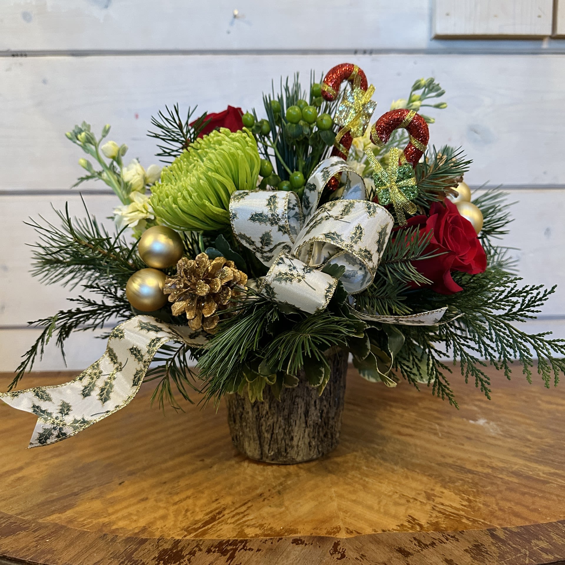 A Special Gift - Red roses, yellow stock, green chrysanthemums, and green hypericum arranged in a log pot with conifer greens, a seasonal holly bow, gold pinecones, gold ornament balls, and candy cane present inserts.