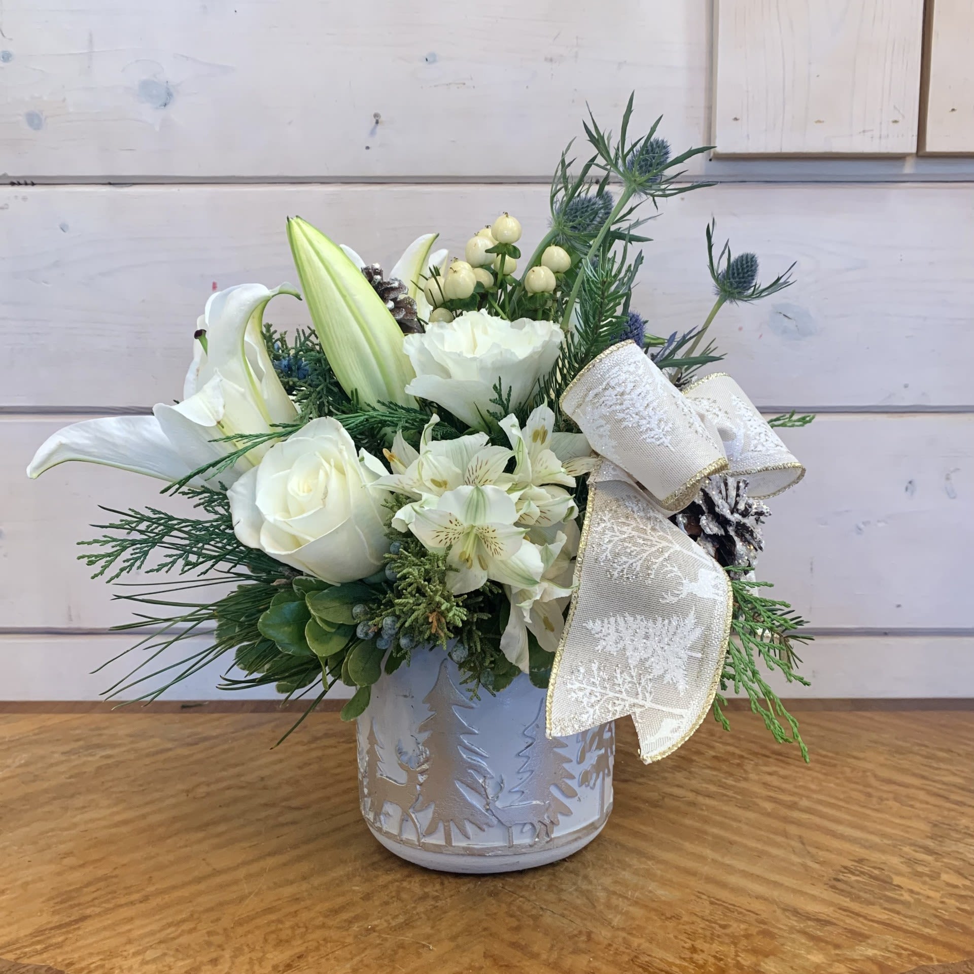 A Winter's Day Bouquet - A wintry arrangement of white lilies, white roses, white lisianthus and white hypericum paired with blue thistle in a silver reindeer motif pot with white alstroemeria, a winter tree bow and snowy iridescent pinecones. Approximately 12&quot; tall and 8&quot; wide.