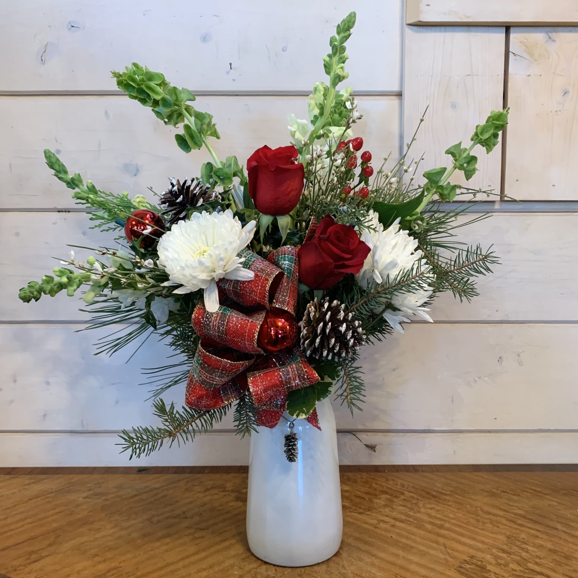 All I Want For Christmas - All I want for Christmas is you... and this arrangement of red roses, white snapdragons, bells of ireland, and white chrysanthemums with red hypericum berries in a tall white vase with an elegant pinecone charm with seasonal filler flower, a sparkly plaid bow, iridescent pinecones and shiny red ornament balls. Approximately 17" tall and 11" wide.