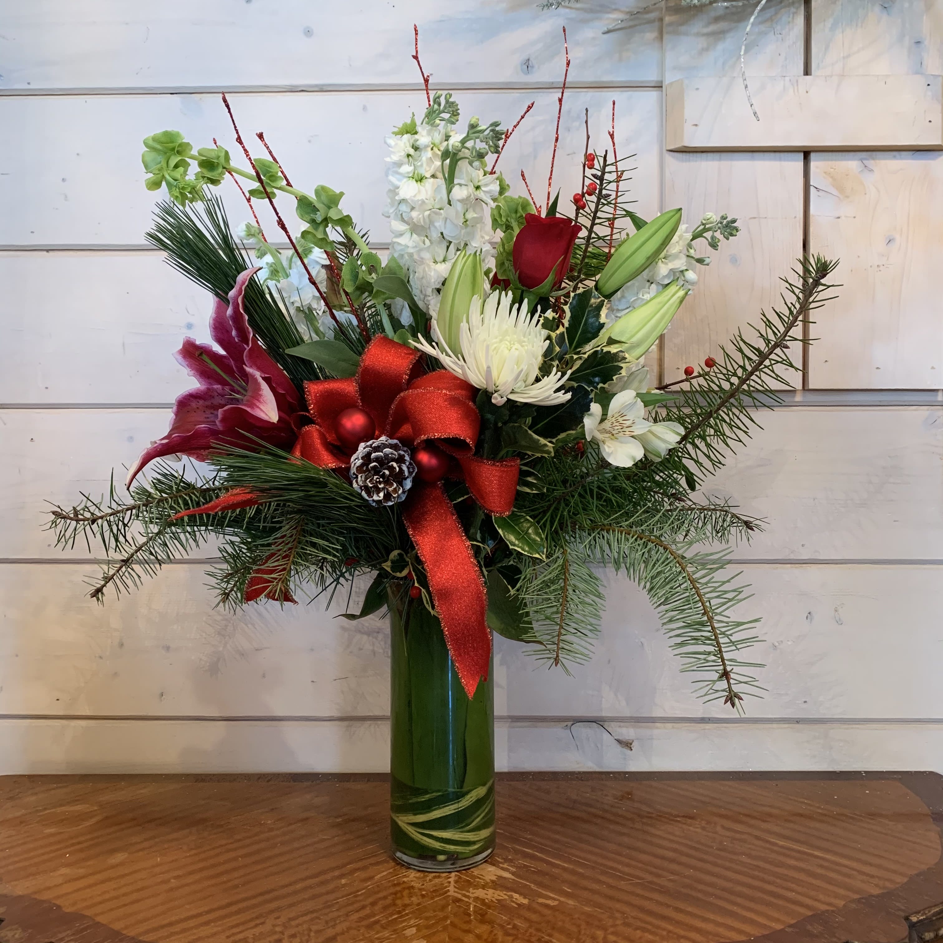 Celebrate the Season  - red roses, burgundy lily, white stock, bells of Ireland, white chrysanthemums, white alstromeria arranged in a 9in leaf lined cylinder with seasonal evergreens, , red glitter branches, red berries, large red bow, pinecones and red ornament balls. Approximately 20in tall and 15in wide. 