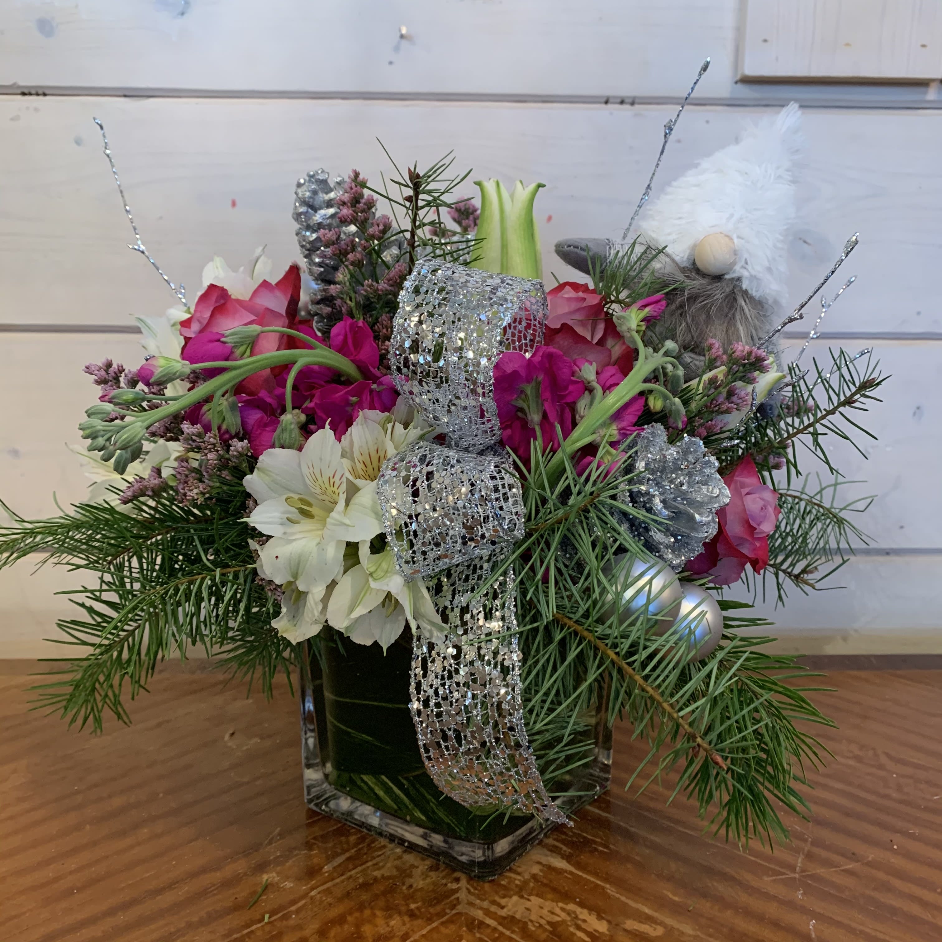 December Magic  - lavender roses, magenta stock, white lily, white alstromeria, and purple misty arranged in a leaf lined 5in cube with seasonal evergreens, silver glitter branches, a sliver bow, silver pinecones and ornament balls. Includes a small white and grey gnome. approximately 12in tall and 12in wide.  