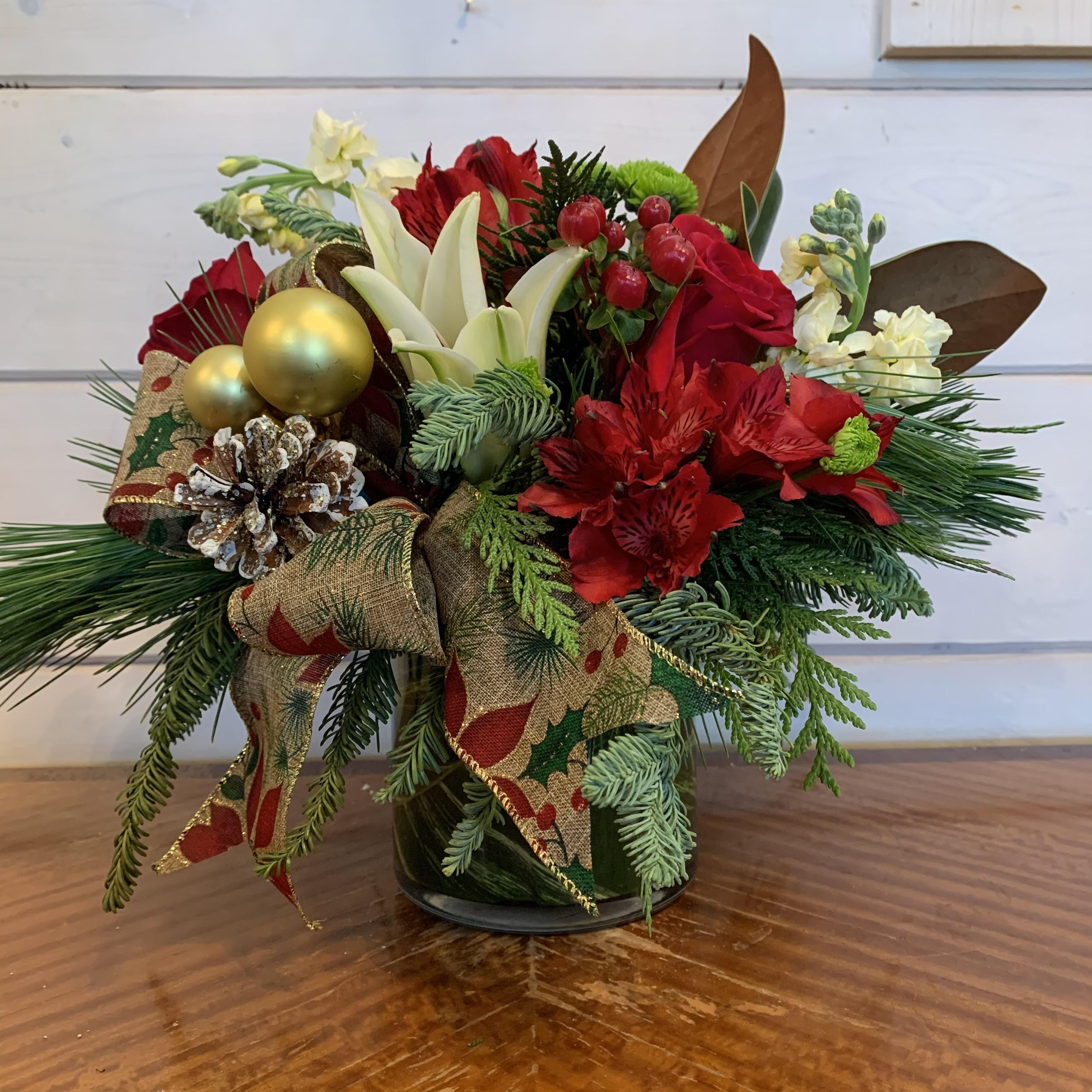 Deck the Halls - Red roses, white lily, yellow stock, green buttons and red alstromeria arranged in a medium leaf lined round vase with Christmas greens, magnolia, a festive bow, pine cone and gold ornament balls. Approximately 12in tall and 12in wide. 