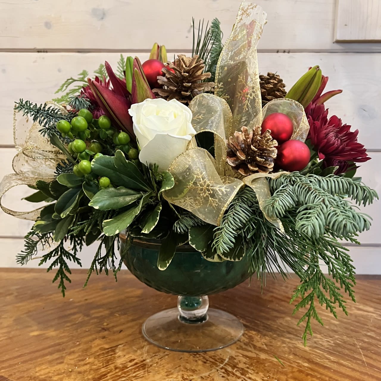 Elegant Holiday Centerpiece  - White roses, burgundy lilies, green hypericum berries, and burgundy chrysanthemums arranged in a cut glass pedestal bowl with seasonal evergreens, large festive gold bows, gold pinecones and red ornament balls. Approximately 14in tall and 16in wide.  
