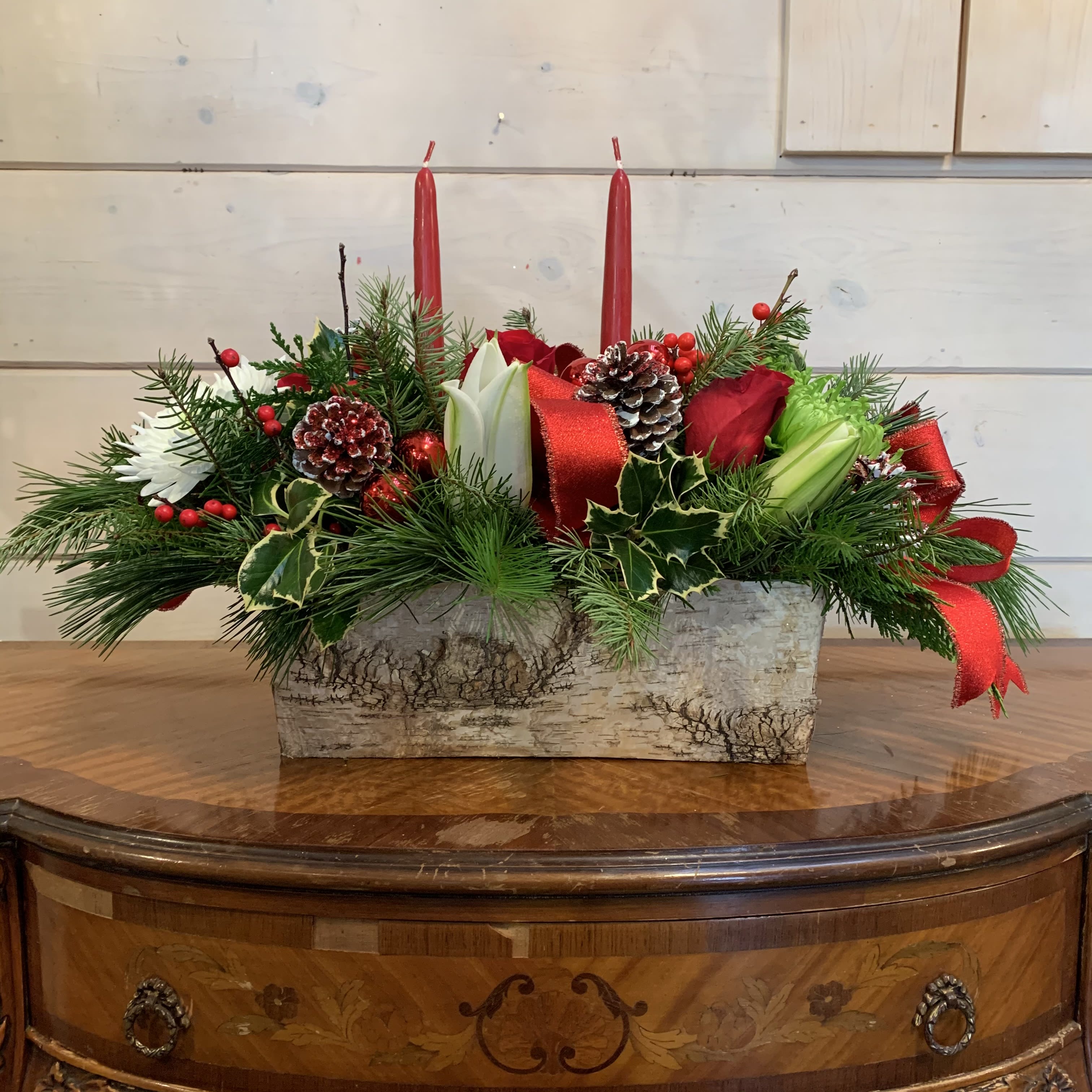 Elaine's Favorite Centerpiece  - Red roses, white lilies, green and white chrysanthemums, and ilex berries arranged in a long and low birch box with seasonal evergreens, red ribbon, pinecones, red ornament balls and to red candles. Approximately 20in long and 10in tall. 