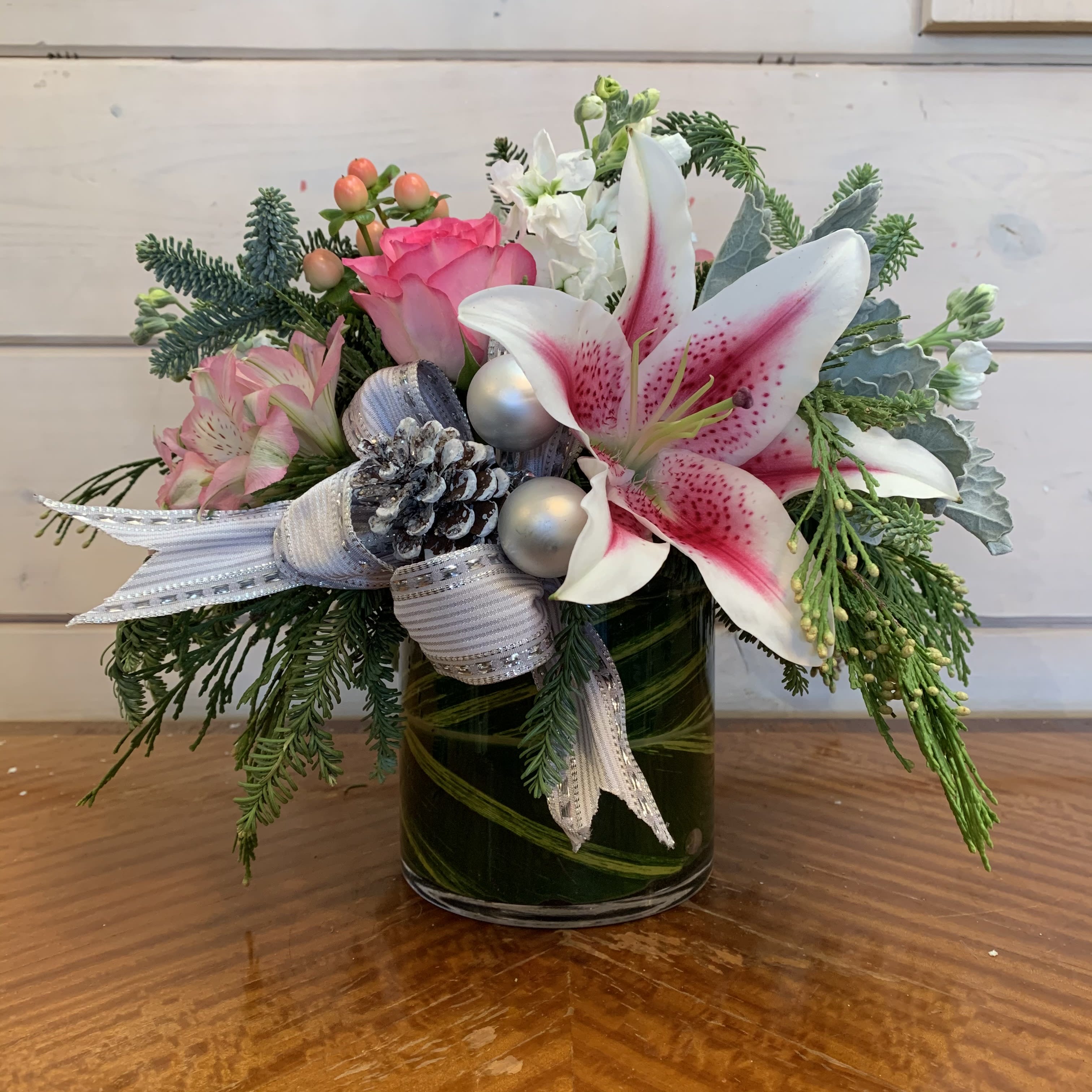 Fun and Frosty  - Pink roses, stargazer lilies, pink hypericum berries, white stock and pink alstromeria arranged in a medium leaf lined round vase with Christmas greens, dusty miller, and a festive silver bow, pinecone and silver ornament balls. Approximately 11in tall and 11in wide. 