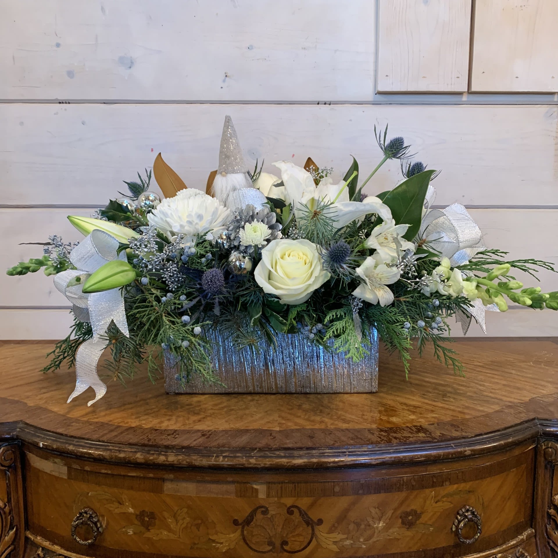 Ginger's Favorite Centerpiece - This whimsical wintry centerpiece feautures a little gnome along with white roses, white lilies, white chrysanthemums, white snapdragons, and blue thistle in silver rectangular planter with white alstroemeria, magnolia branches, and silver accents of seeded eucalypus, pinecones, ornament balls and bows. Approximately 15" long for the standard, 17" long for the deluxe, and 20" long for the premium (in a longer vase).