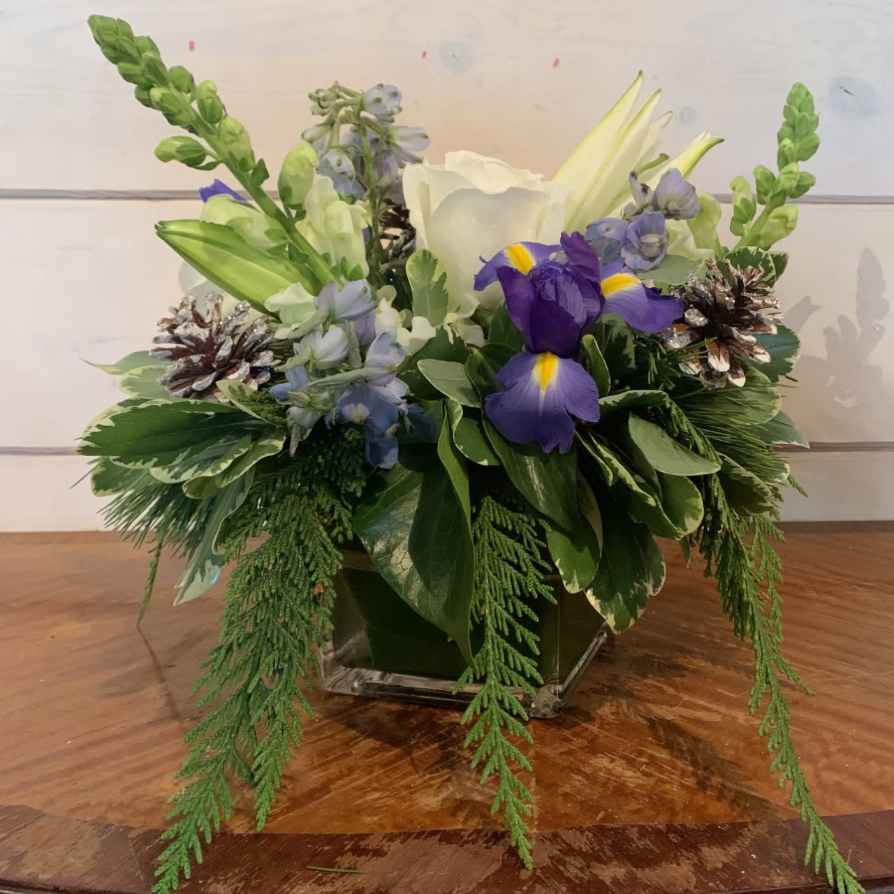 Frosty Winter Pine Bouquet - Mix of blue and white flowers including roses, delphinium, lilies, snapdragons and iris arranged in a medium leaf lined cube vase with winter evergreens and frosted pinecones. Approximately 12in tall and 12in wide. 