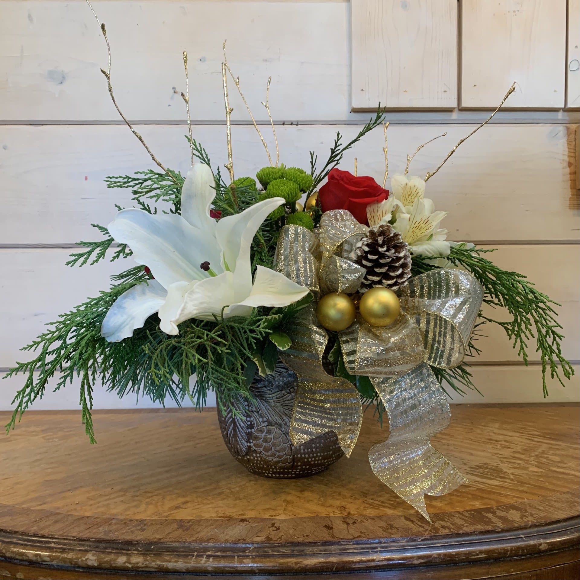 Christmas Tradition - Red roses, white lilies, green buttons, and white alstroemeria arranged in a bronze colored pot with pine motif, with a large striped gold and silver bow, glitter pinecones, gold ornament balls, and gold glitter branches. Approximately 11&quot; wide and 11&quot; tall.