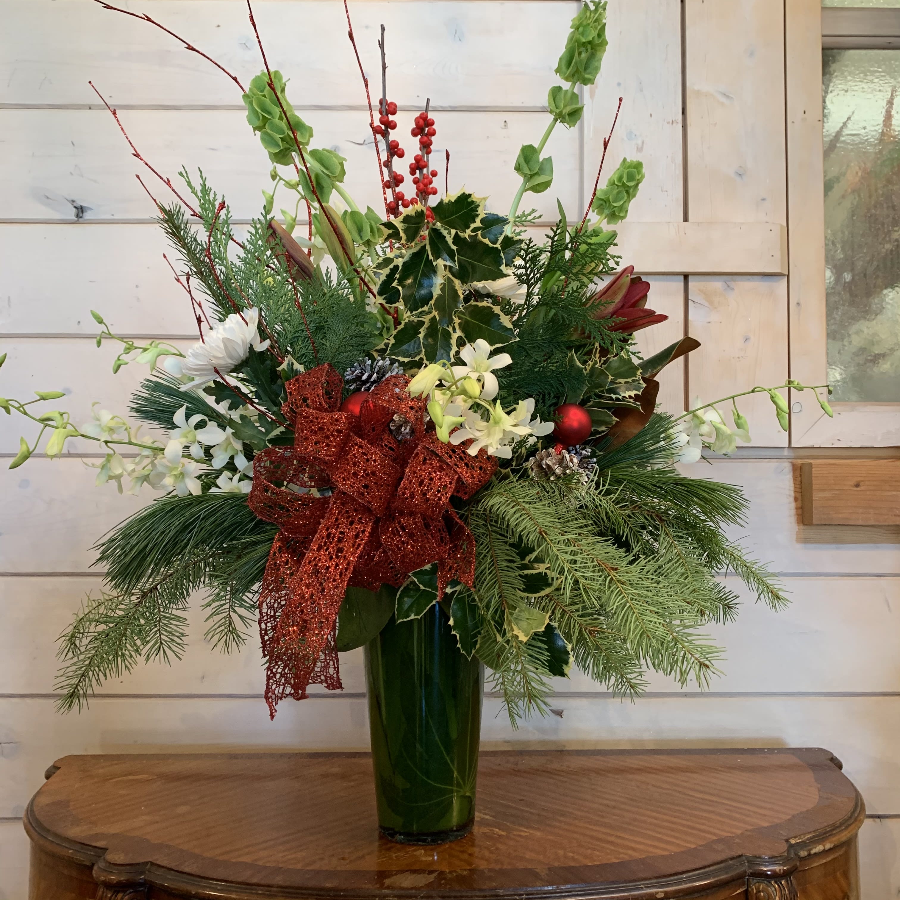 Holiday Blessings  - Burgundy lilies, white orchids, bells of Ireland, white chrysanthemums, and magnolia arranged in a large leaf lined French vase with seasonal evergreens, holly, red berries and red glitter branches. Decorated with a large festive read bow, ornament balls and pinecones. Approximately 28in tall and 22 in wide.  