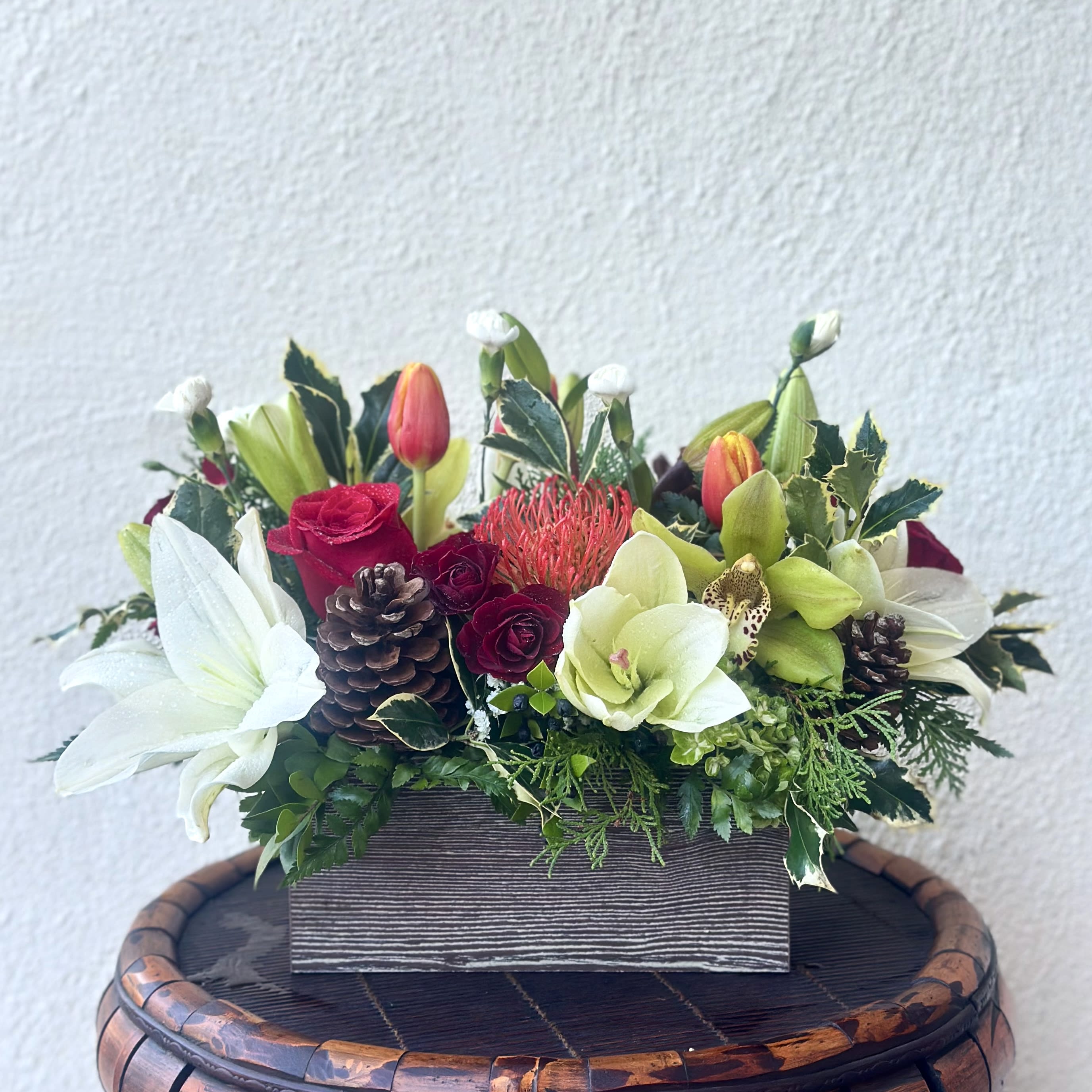 Holiday Box - Celebrate the holidays with this festive centerpiece bouquet on your Tablescape! Designed with a mix of roses, spray roses, lilies, orchids  and tulips with hollies and pinecones in a keepsake rectangular wood box.   Perfect gift not only for Christmas but on any occasion your loved ones are celebrating for this month.   Approximately 12” W x 9” H