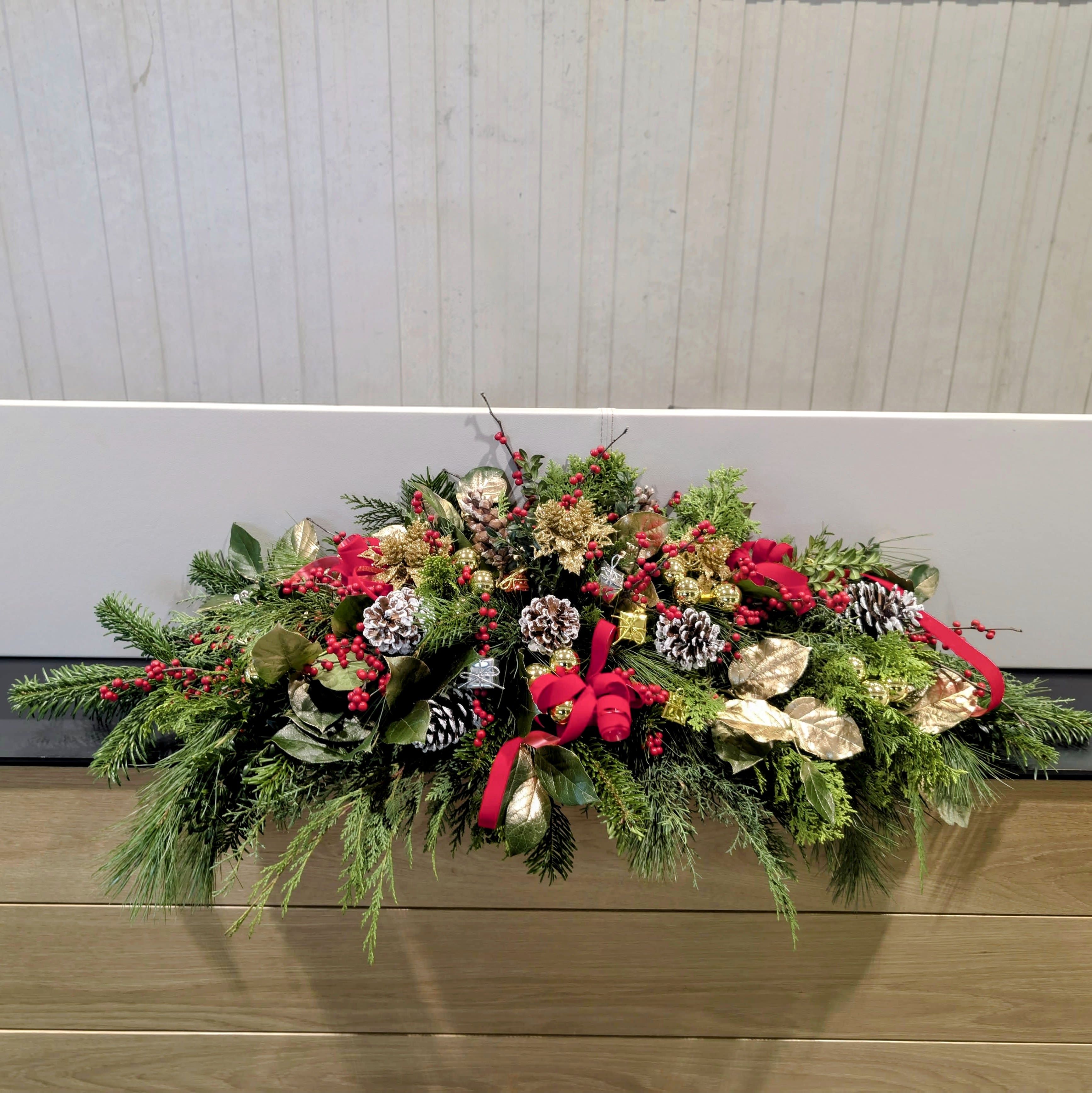 Holiday Splendor Mantlepiece - Deck your mantle or holiday table with this lush and elegant long-and-low centerpiece! Featuring fragrant evergreens, gilded accents, vibrant red berries, pinecones, and festive red ribbons, this arrangement captures the magic of the season. Touches of gold and a harmonious mix of textures make it a stunning statement piece for holiday gatherings or a cozy night by the fire.  Perfect for mantles, dining tables, or as a special gift, this holiday arrangement brings warmth and festive cheer to any space! (Approximately 2 and a half feet)  Pro tip: Let us know if this piece will be used on a mantle or as a centerpiece so we can design it to be one-sided or all-around for the best display!