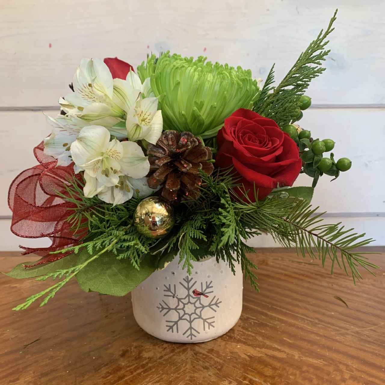 Holiday Wishes Bouquet  - red roses, green chrysanthemums, green hypericum berries and white alstroemeria arranged in a small white ceramic pot with a snowflake and cardinal print. Includes winter evergreens, red bow, pinecones and gold ornament balls. Approximately 10in tall and 10in wide.