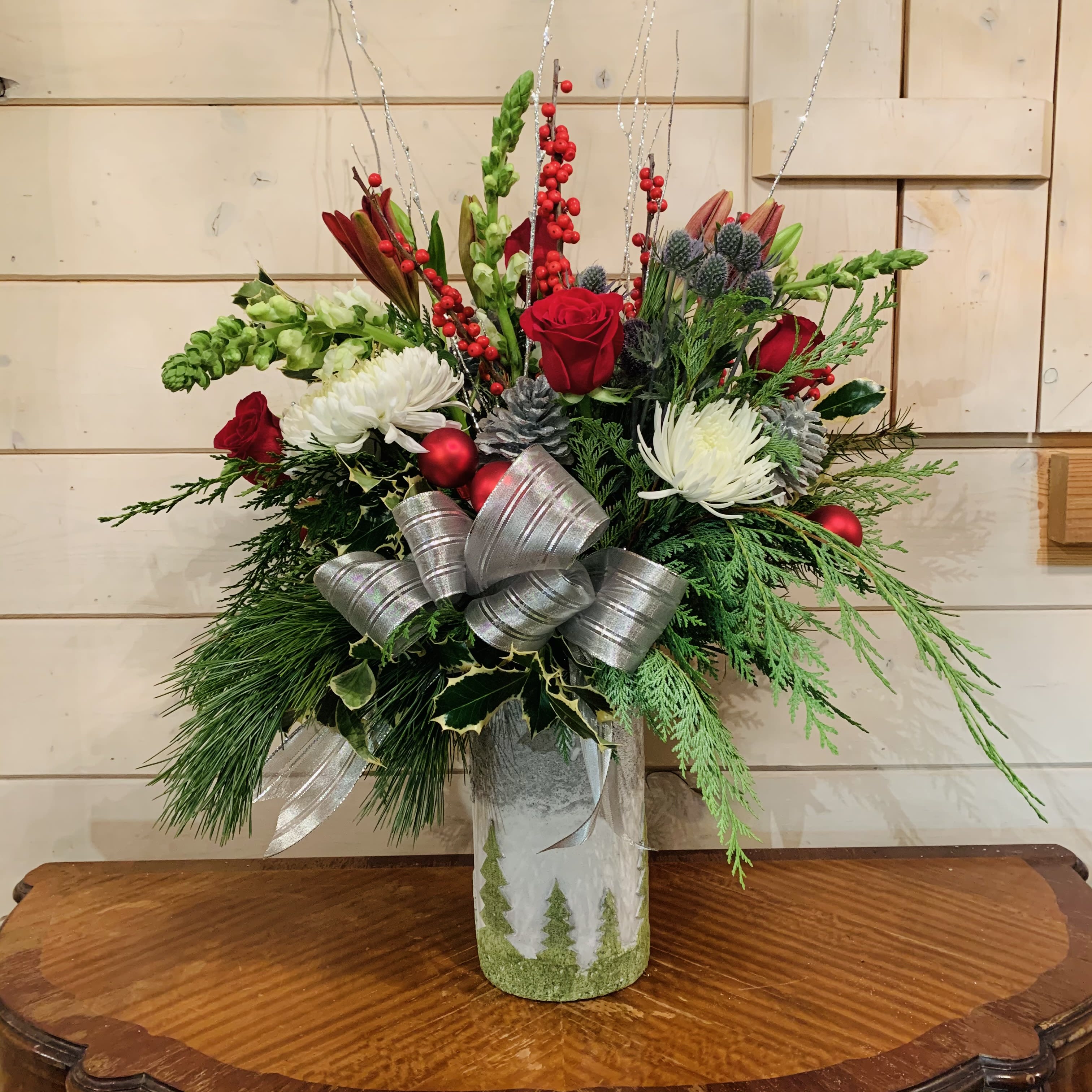 Joy of the Season  - Red roses, burgundy lily, ilex berries, white snapdragons, blue thistle, white chrysanthemums and holly branches arranged in a large textured wide 9in cylinder ceramic vase with tree design. Decorated with Silver glitter branches, a large silver bow, silver glitter pinecones and red ornament balls. Approximately 24in tall and 18in wide.  