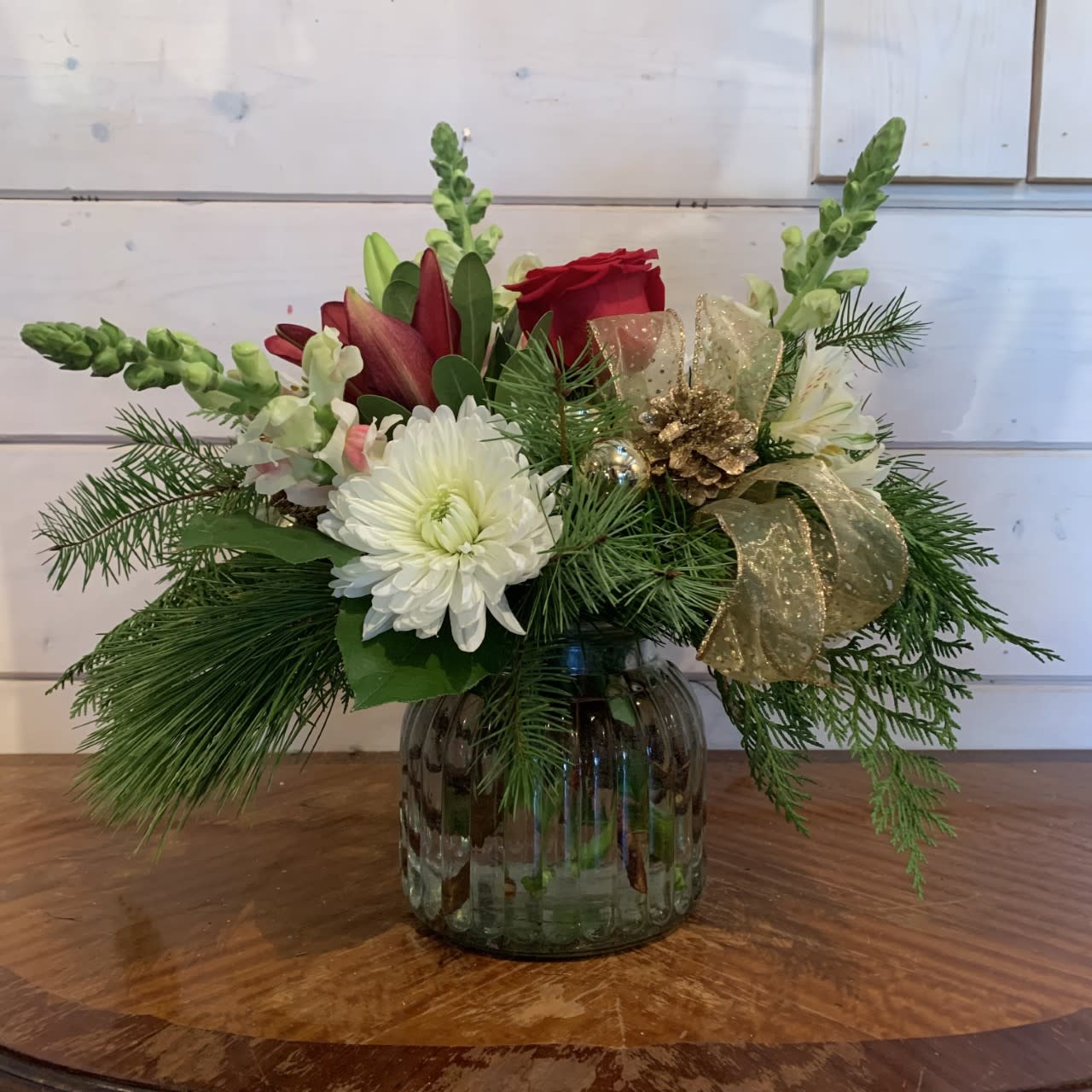 Happy Holidays  - red roses, burgundy lily, white snapdragons, white chrysanthemums and alstroemeria arranged in a large ribbed glass jar with winter evergreens gold bow, gold pinecones, and gold ornament balls. Approximately 12in tall and 12in wide.   