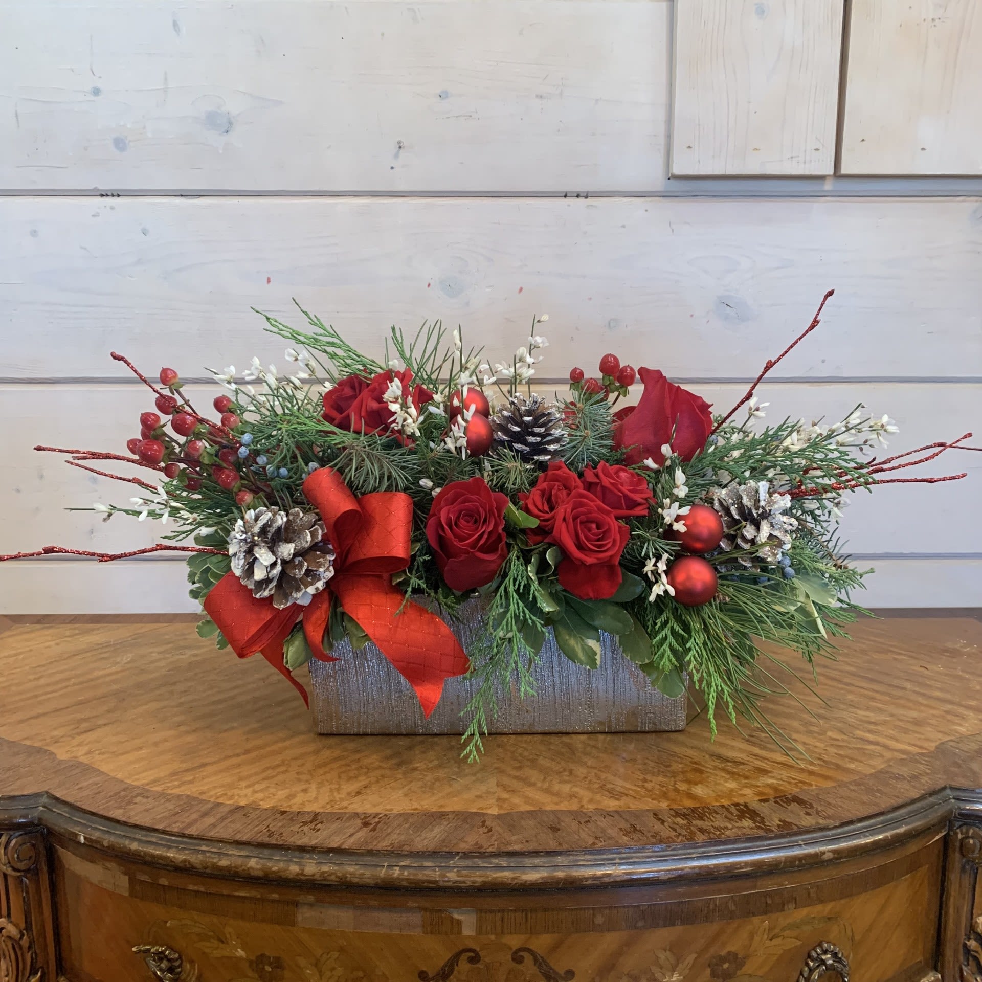 Lakeitha's Favorite Centerpiece - A traditional christmas centerpiece of red roses, red spray roses, and red hypericum in a silver rectangle with white ginestra, red bows, snowy iridescent cones, red ornament balls, and red glitter branches. Approximately 14&quot; long for the standard, 16&quot; long for the deluxe and 20&quot; long for the premium.